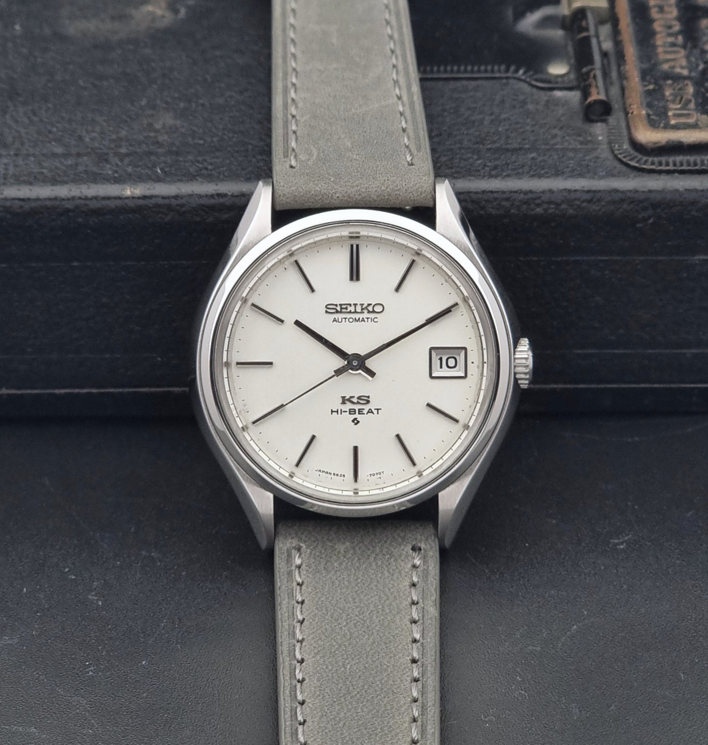 A Vintage King Seiko 5625-7121 watch with a silver-toned metal case, white dial, and a light gray leather strap, displayed against a dark textured background.