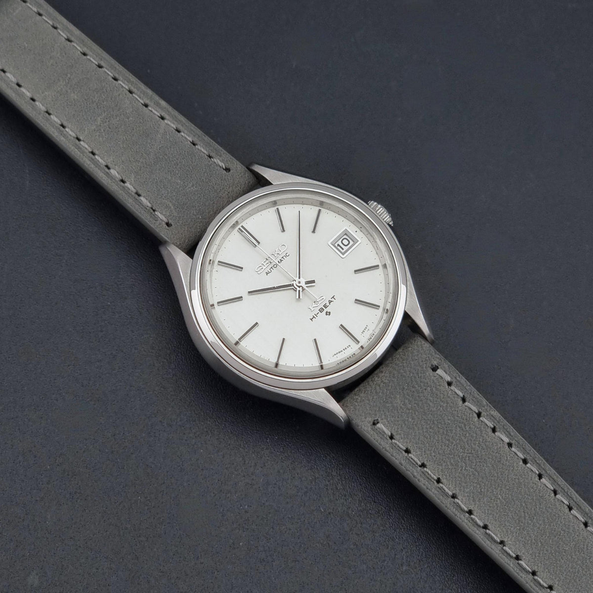 A Vintage King Seiko 5625-7121 watch with a silver-toned metal case, white dial, and gray leather strap, photographed diagonally on a dark textured background.