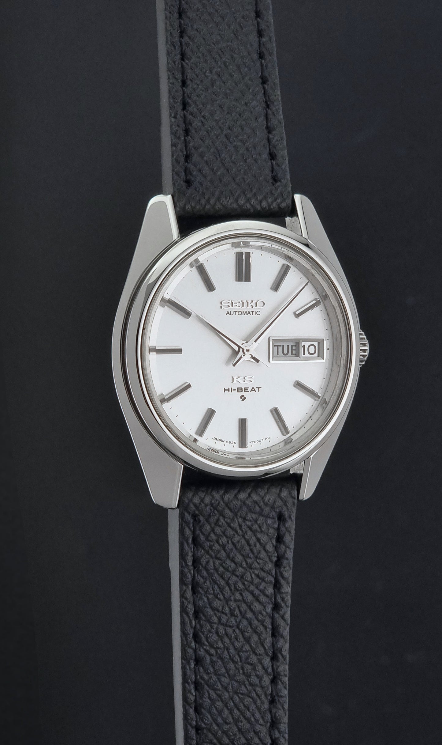 A Vintage King Seiko Wristwatch 5626-7000 automatic watch with a silver-toned metal case, white dial, and textured black strap, shown against a dark background.