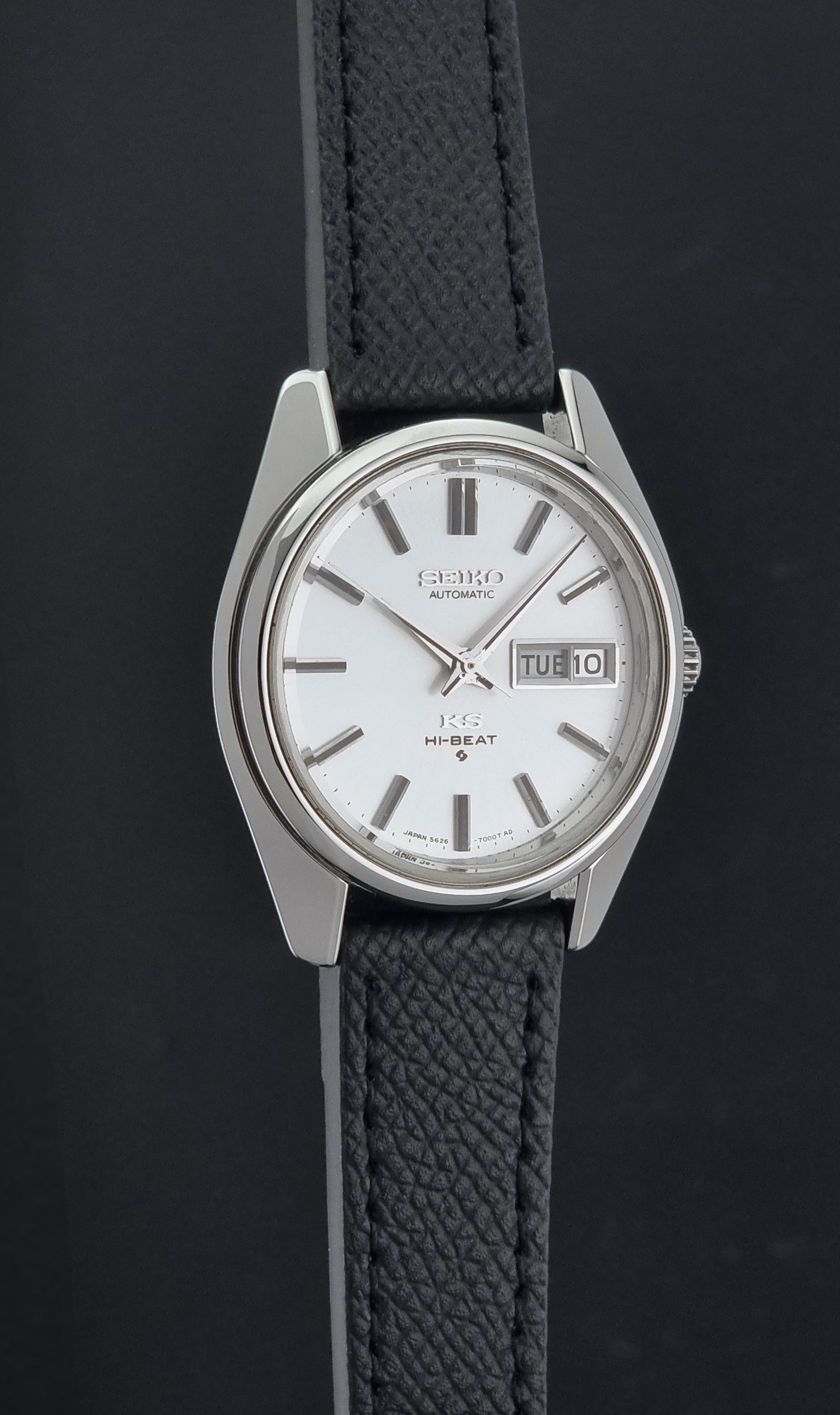A Vintage King Seiko Wristwatch 5626-7000 automatic watch with a silver-toned metal case, white dial, and textured black strap, shown against a dark background.