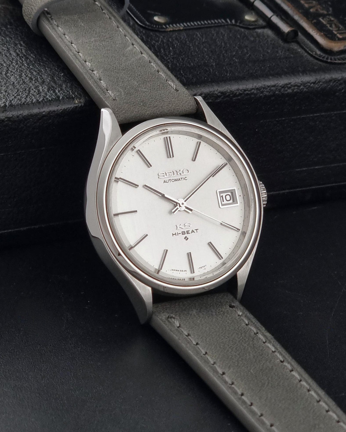 A Vintage King Seiko 5625-7121 watch with a silver-toned case, white dial, and grey leather strap, resting on a black leather surface.