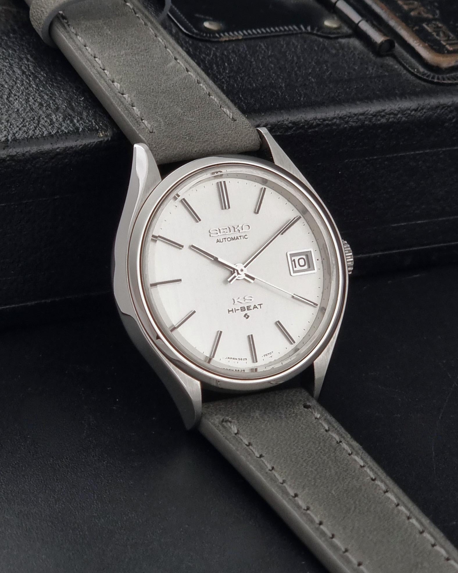 A Vintage King Seiko 5625-7121 watch with a silver-toned case, white dial, and grey leather strap, resting on a black leather surface.
