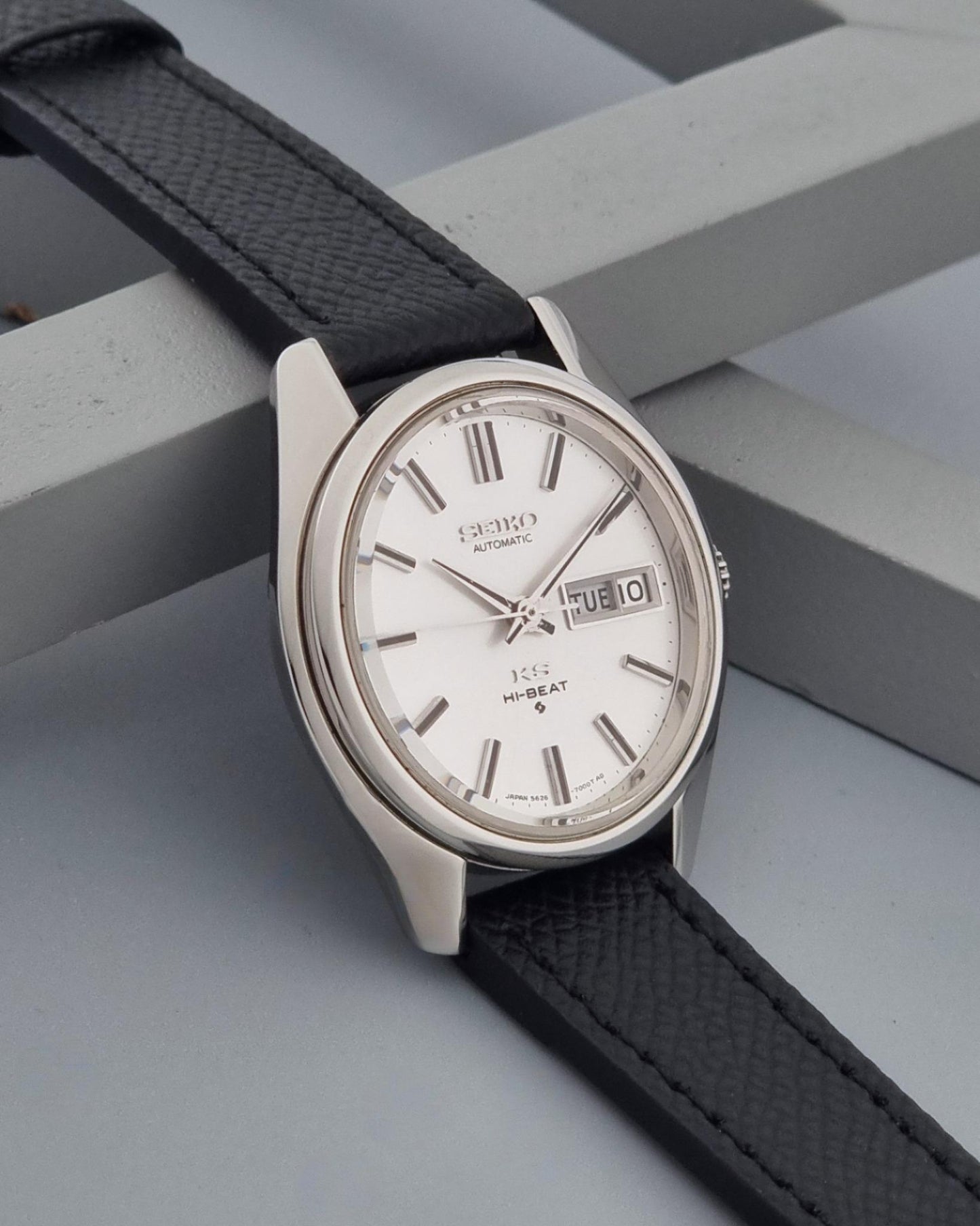 A Vintage King Seiko Wristwatch 5626-7000 with a silver-toned oval case, white dial, and black textured leather strap, resting on a gray geometric surface.