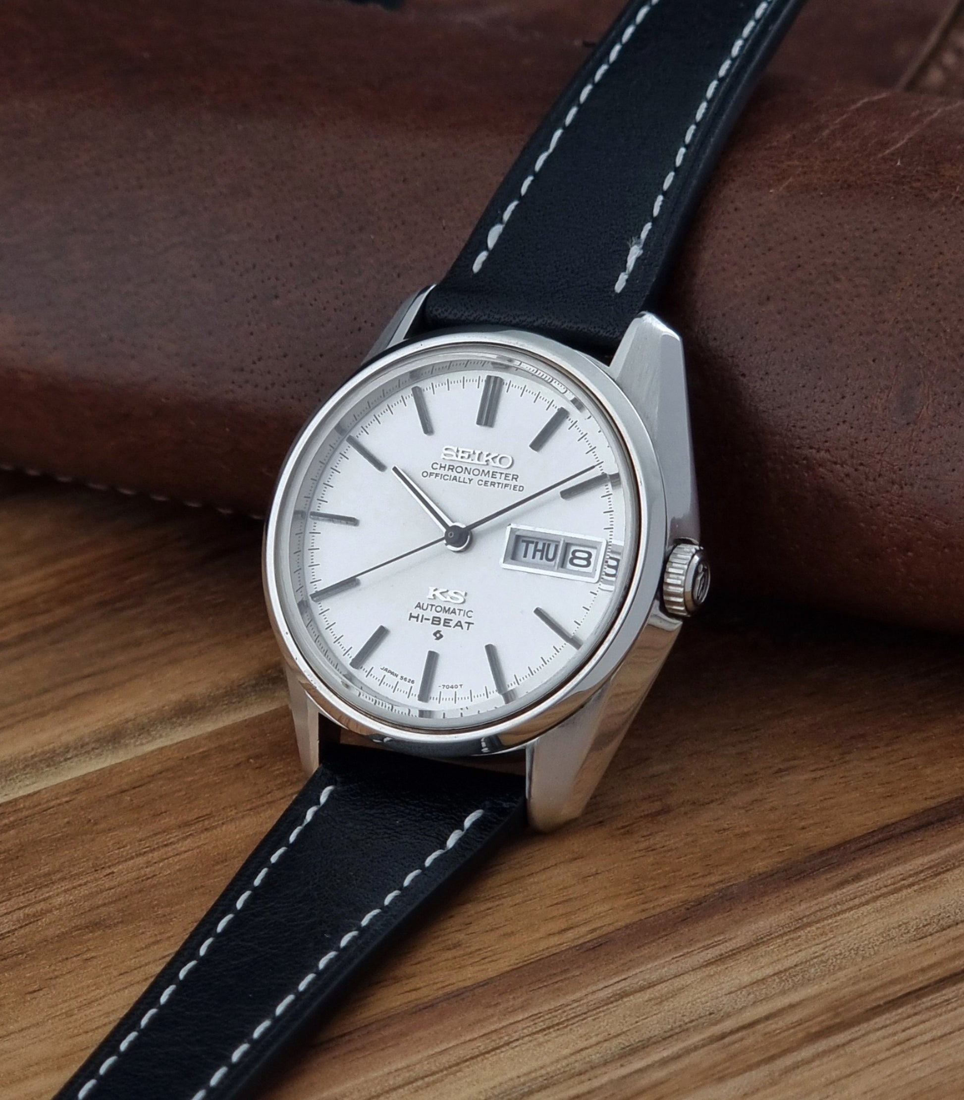 Vintage King Seiko watch 5626-7041  with a silver-toned metal case, white dial, and black leather strap with white stitching, on a wooden surface crown view