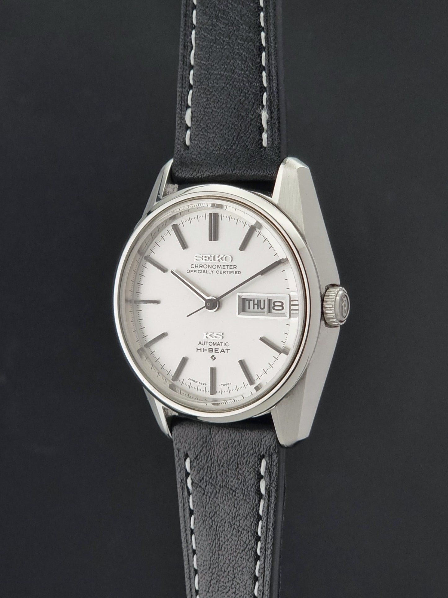 Vintage King Seiko watch 5626-7041  with a silver-toned metal case, white dial, and black leather strap with white stitching, on a dark background crown profile view