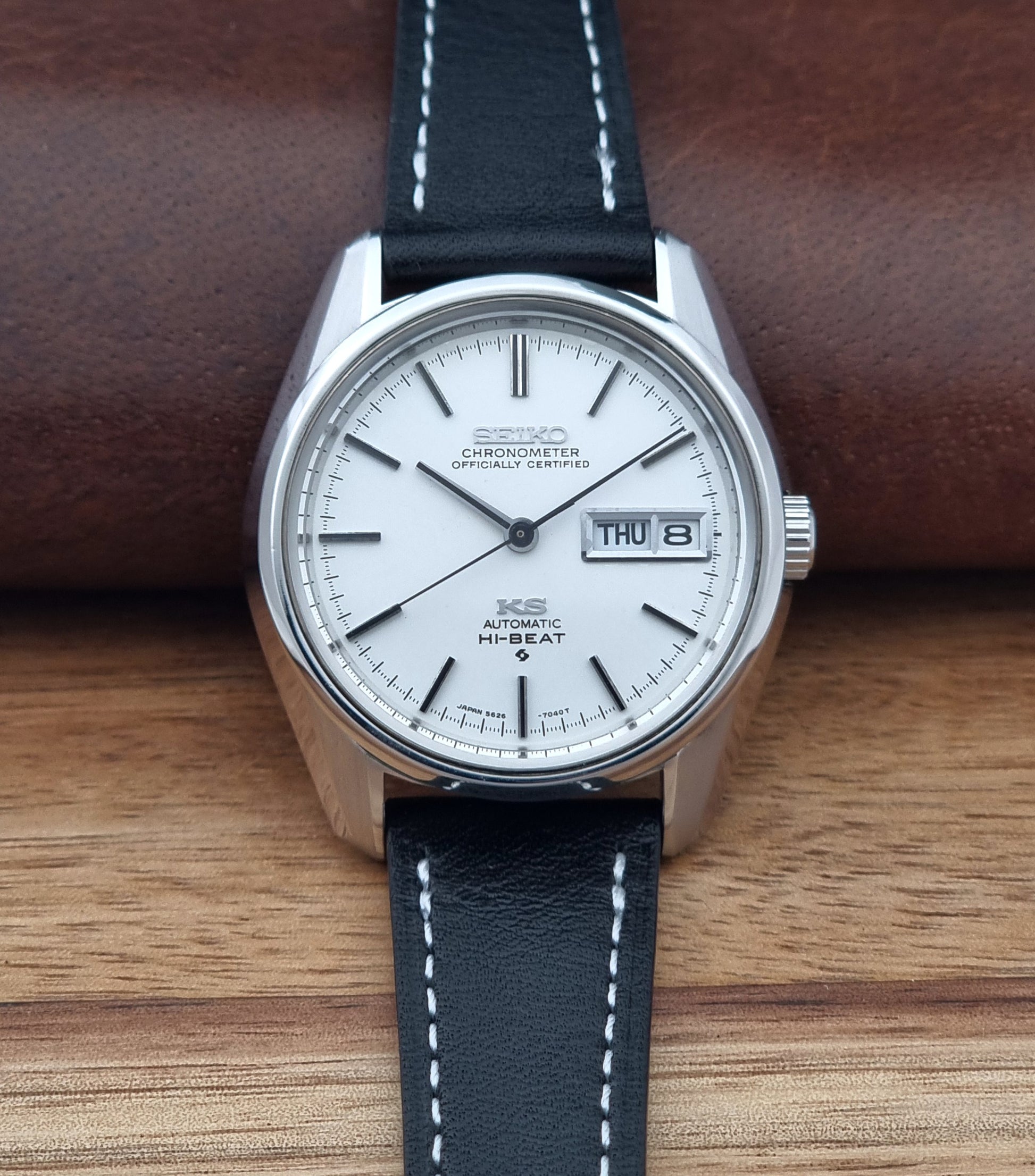 Vintage King Seiko watch 5626-7041  with a silver-toned metal case, white dial, and black leather strap with white stitching, on a wooden surface dial view