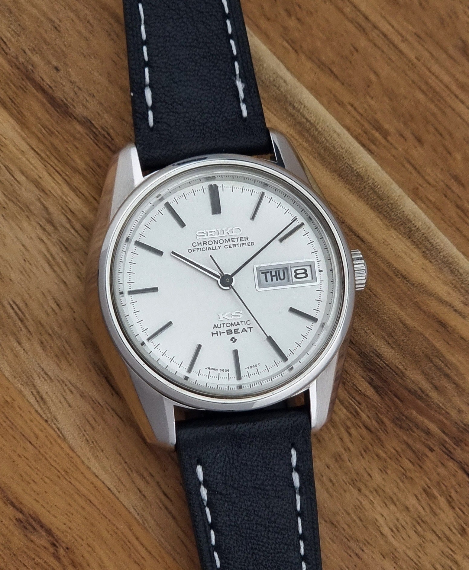 Vintage King Seiko watch 5626-7041  with a silver-toned metal case, white dial, and black leather strap with white stitching, on a wooden surface front view