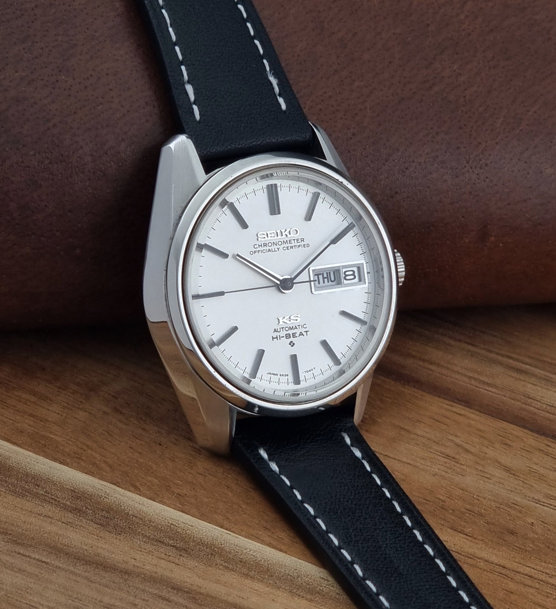 Vintage King Seiko watch 5626-7041  with a silver-toned metal case, white dial, and black leather strap with white stitching, on a wooden surface side view