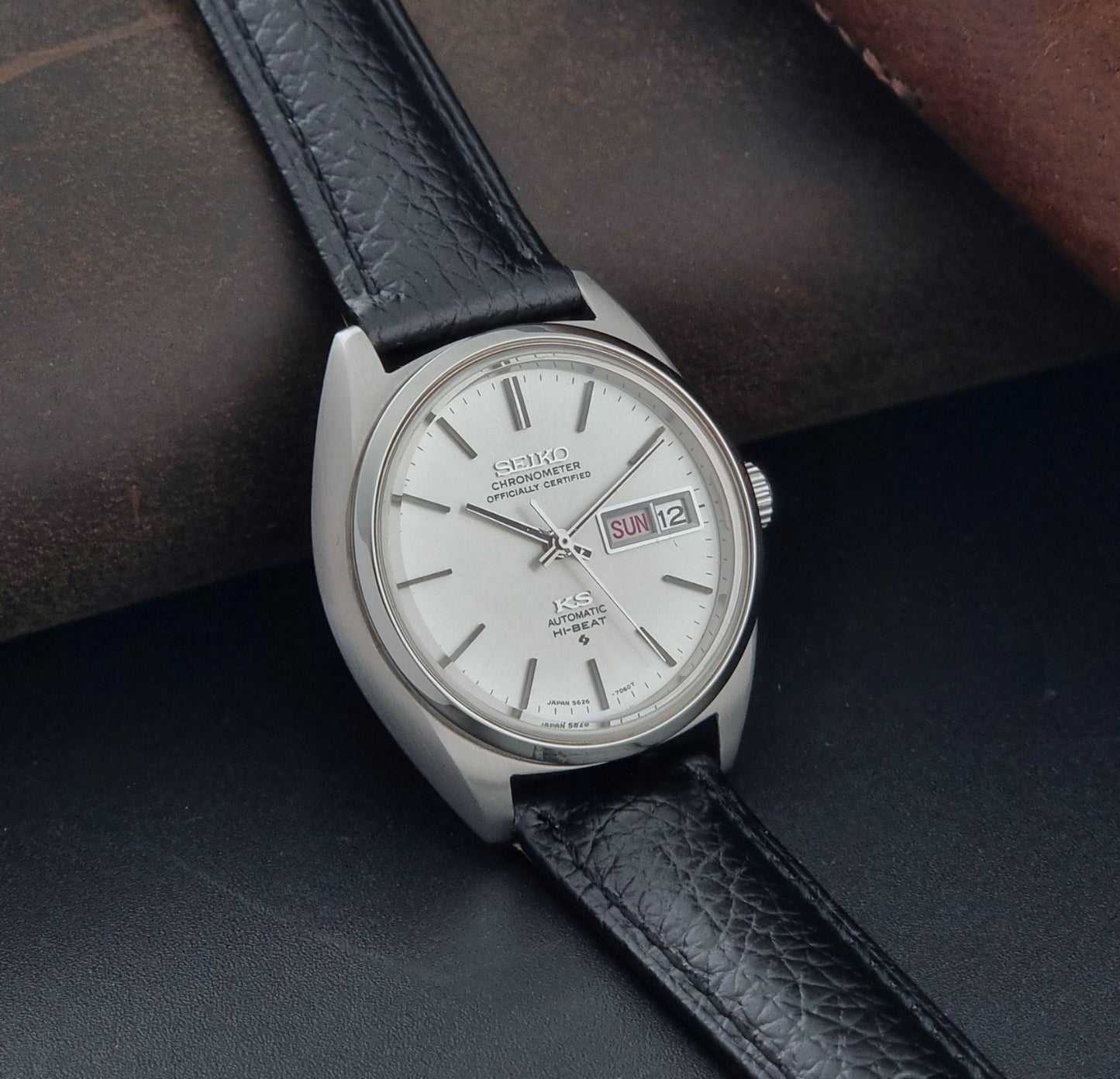 Vintage King Seiko 5626-7060 wristwatch with a silver case and black leather strap on a dark surface side view