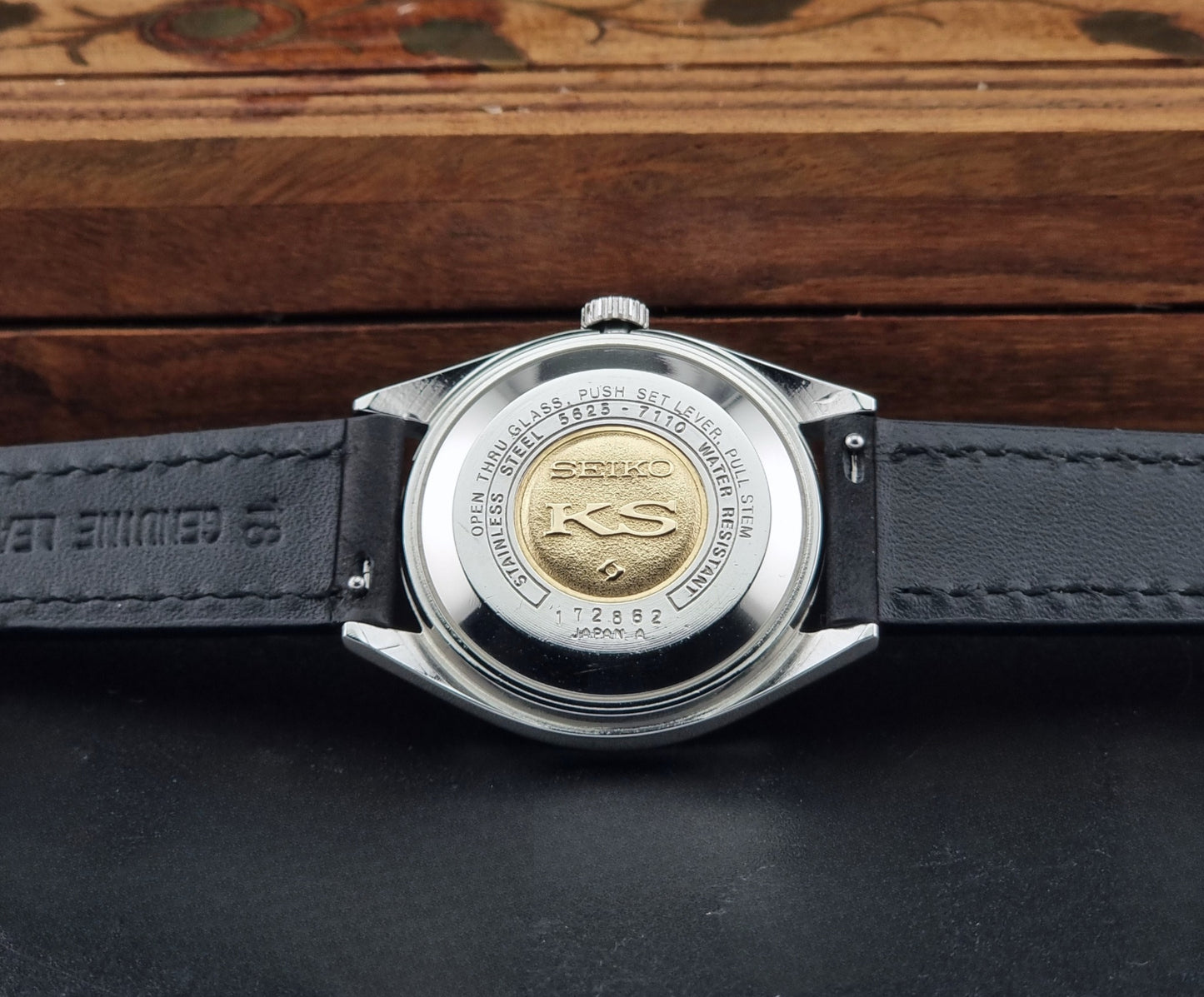 Close-up of the back of a Vintage King Seiko 5625-7110 with a silver stainless steel case and a black leather strap, resting on a dark surface with a wooden box in the background