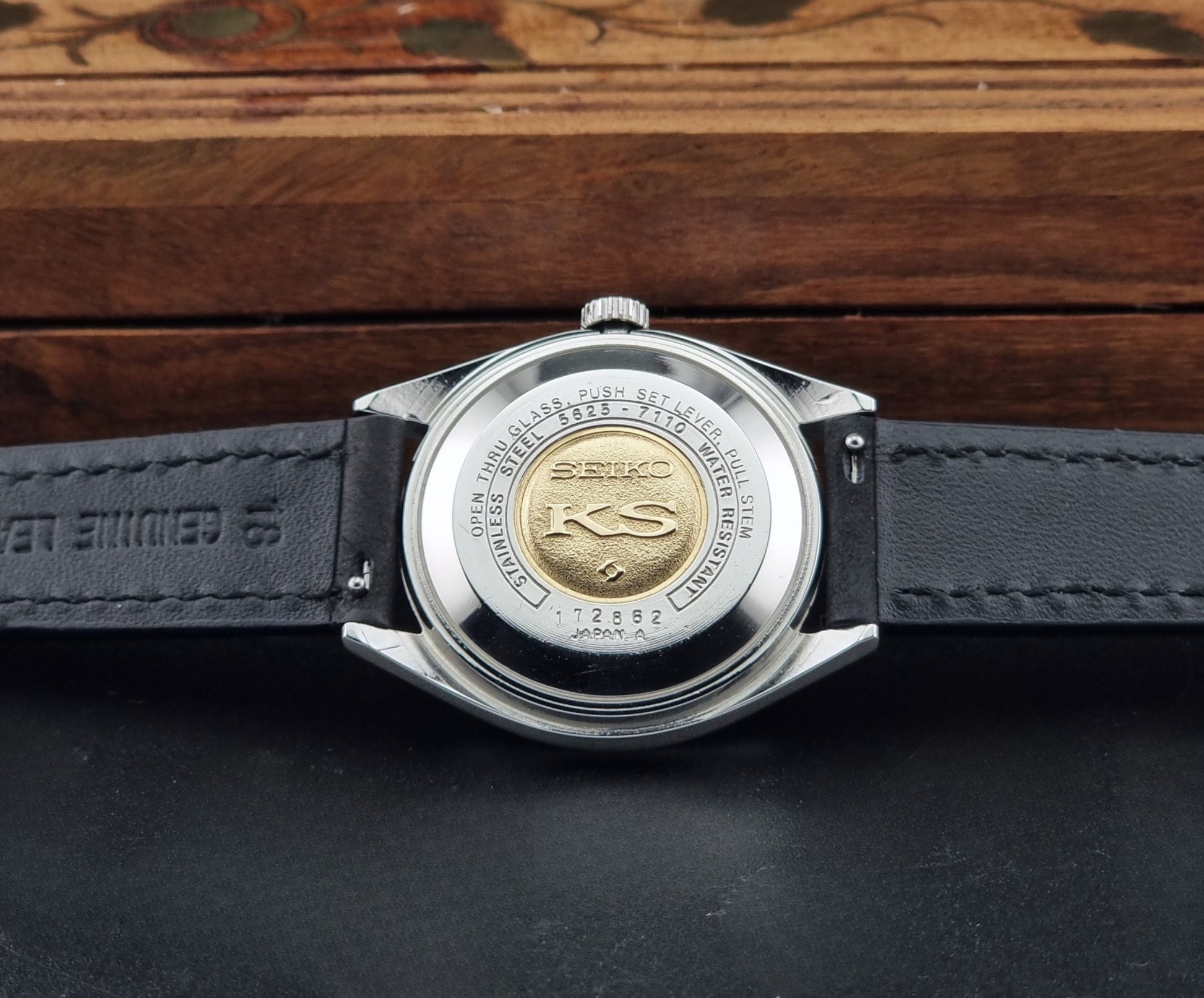 Close-up of the back of a Vintage King Seiko 5625-7110 with a silver stainless steel case and a black leather strap, resting on a dark surface with a wooden box in the background
