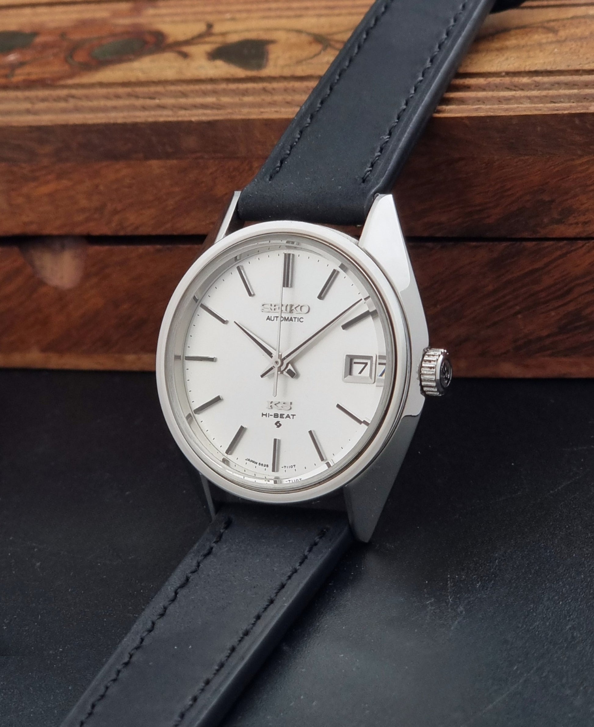 Vintage King Seiko 5625-7110 Wristwatch with a silver-toned metal case, white dial, and black leather strap, on a dark textured background crown view