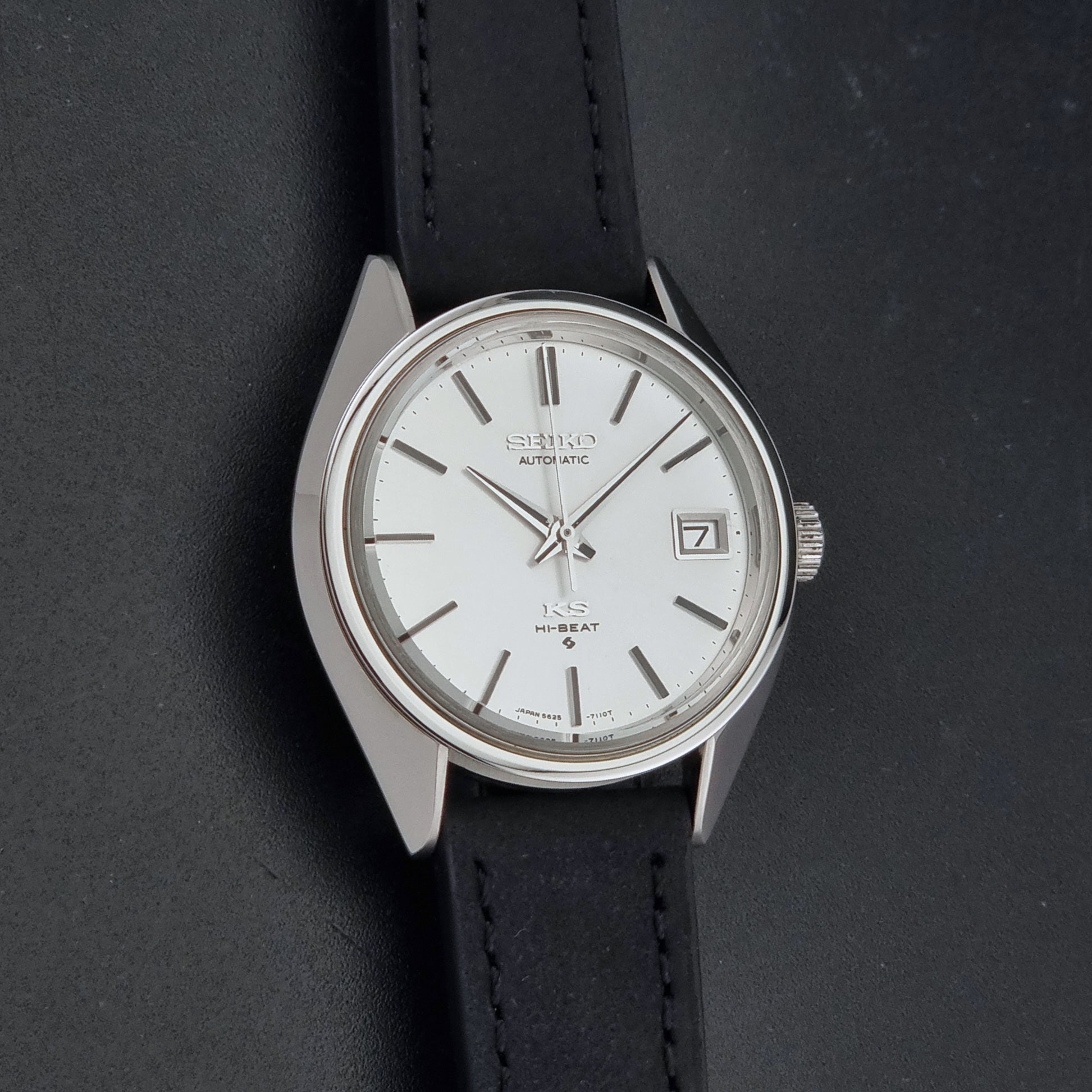 Vintage King Seiko 5625-7110 wristwatch with a silver-toned metal case, white dial, and black leather strap, shown on a dark textured background front view