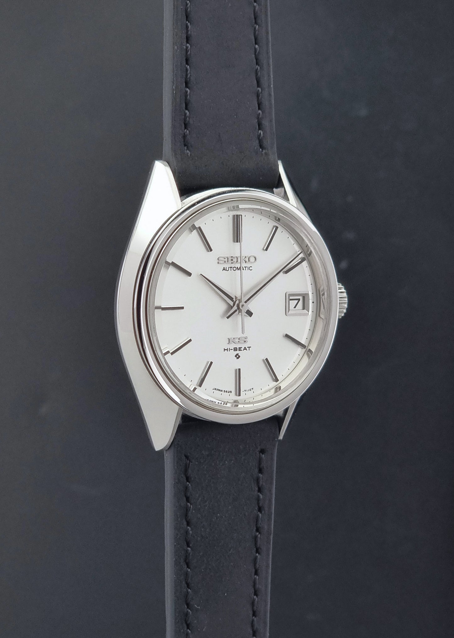 Vintage King Seiko 5625-7110 wristwatch with a silver-tone case, white dial, and black leather strap, shown against a dark gray background sid eprofile view