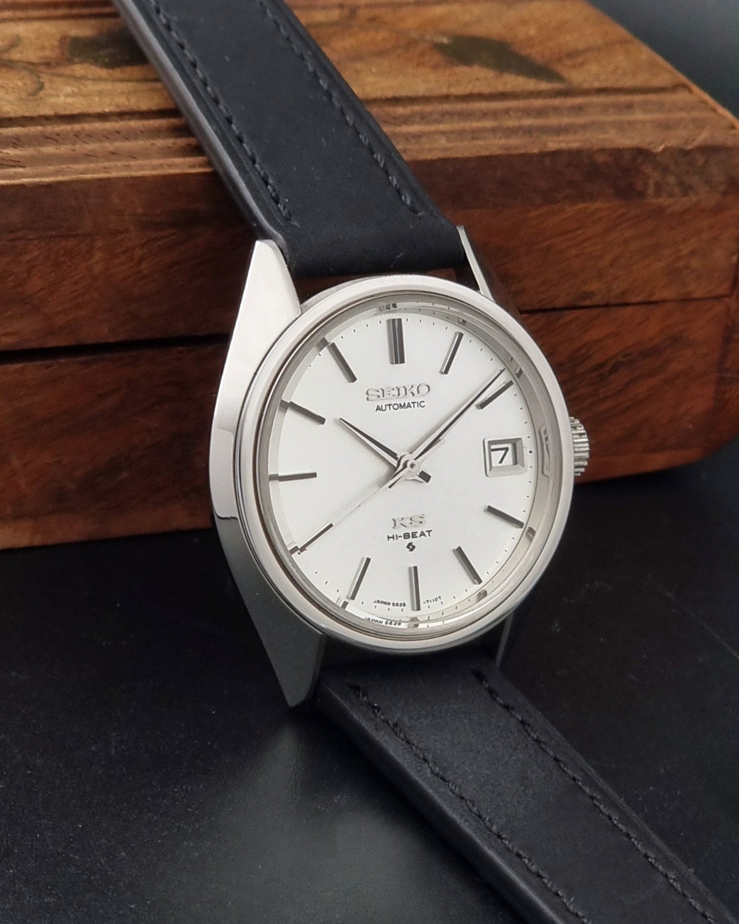 Vintage King Seiko 5625-7110 Wristwatch with a silver-toned metal case, white dial, and black leather strap, on a dark textured background side view
