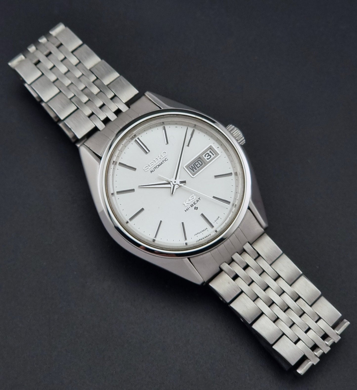 A vintage-style stainless steel wristwatch with a silver-toned metal link bracelet, a white dial, and a date window, photographed on a dark textured background.