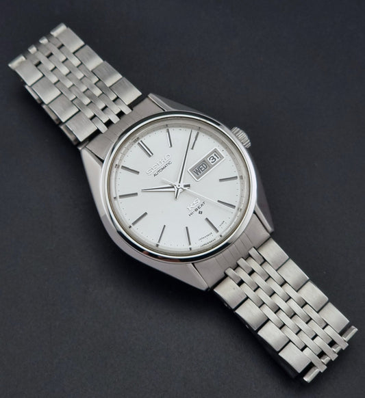 A vintage-style stainless steel wristwatch with a silver-toned metal link bracelet, a white dial, and a date window, photographed on a dark textured background.