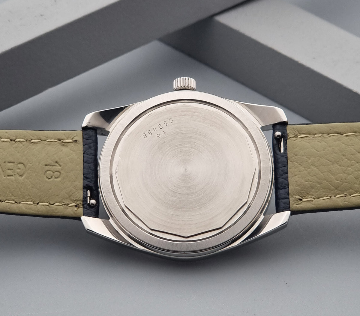Close-up of the back of a Vintage Longines Admiral HF 2302 watch with a brushed silver-tone metal case and a leather strap, photographed on a gray geometric surface.