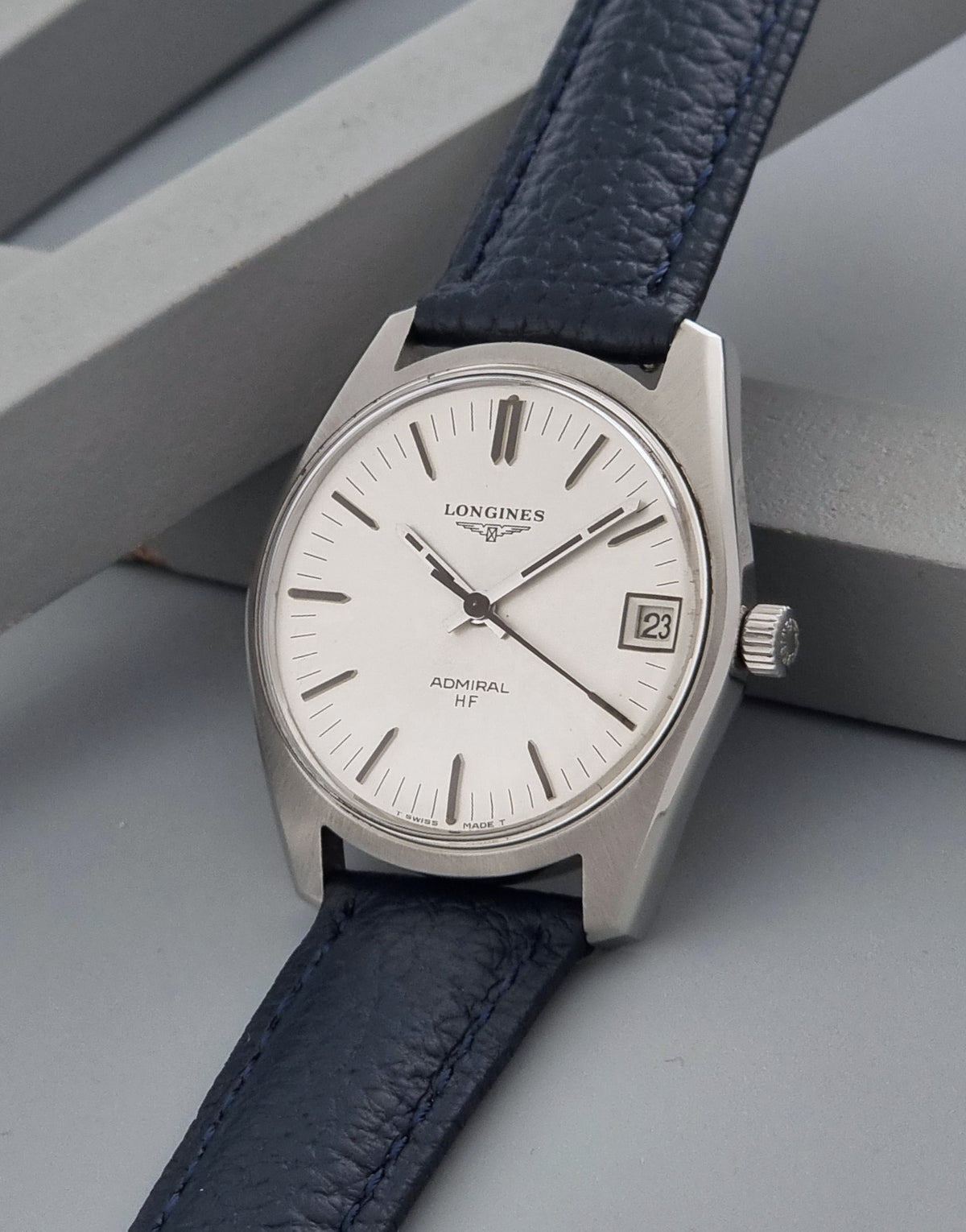 A Longines Admiral HF wristwatch with a silver-toned stainless steel case, white dial, and dark navy blue leather strap, resting on a gray geometric surface.