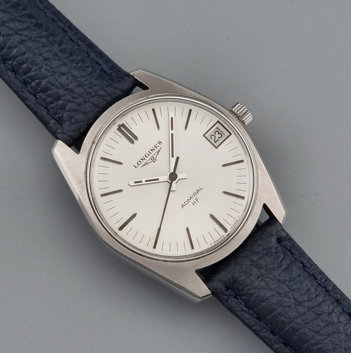 Vintage Longines Admiral HF 2302 watch with a silver-toned metal case, white dial, and dark blue textured leather strap, photographed on a plain gray background.