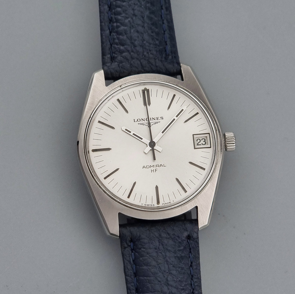 A vintage Longines Admiral HF wristwatch with a silver-toned metal case, white dial, and textured dark blue leather strap, shown against a neutral gray background.