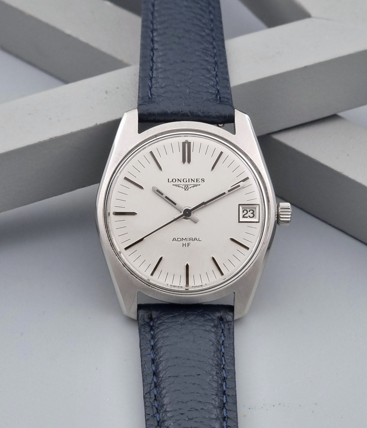 A vintage Longines Admiral HF wristwatch with a silver-toned stainless steel case, white dial, and a textured blue leather strap, displayed on a gray geometric background.