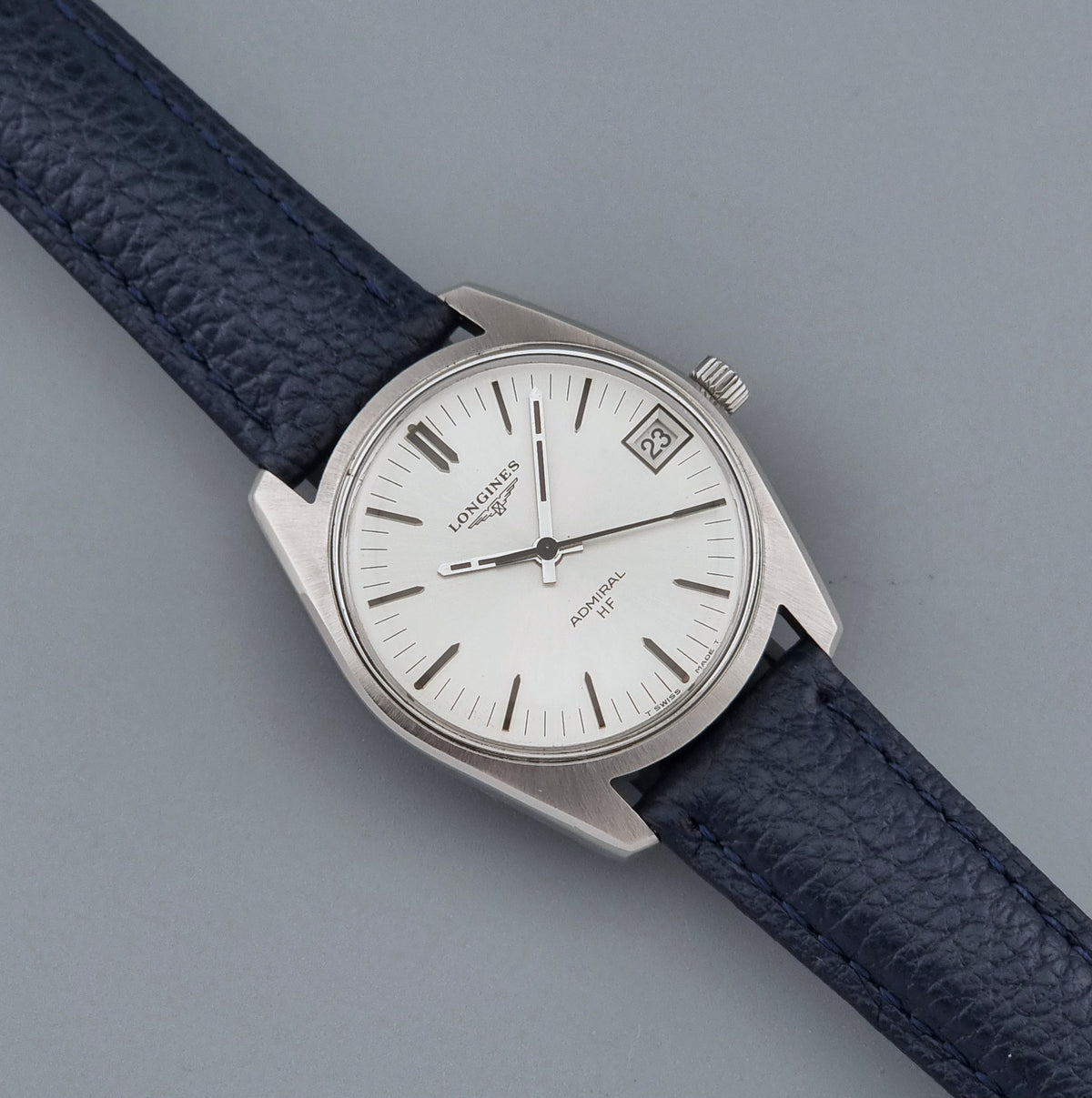 Vintage Longines Admiral HF 2302 watch with a silver-toned metal case, white dial, and dark navy blue textured leather strap, photographed diagonally on a neutral gray background.