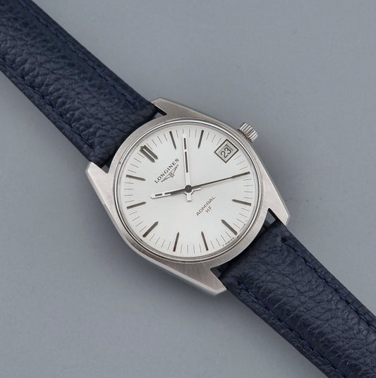 Vintage Longines Admiral HF 2302 watch with a silver-toned metal case, white dial, and dark navy blue textured leather strap, photographed diagonally on a neutral gray background.
