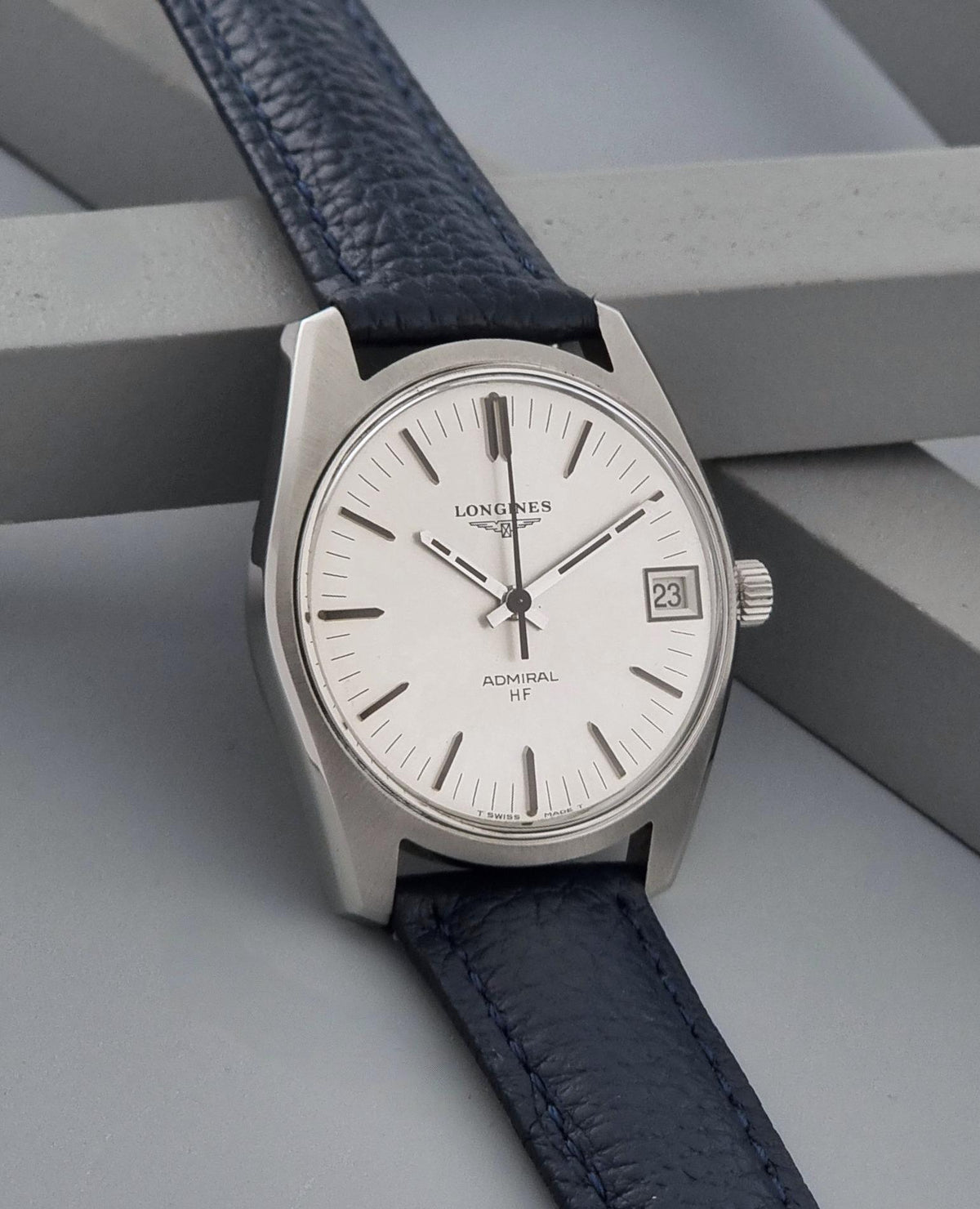 A Longines Admiral HF wristwatch with a silver-toned stainless steel case, white dial, and dark navy blue textured leather strap, resting on a gray geometric surface.