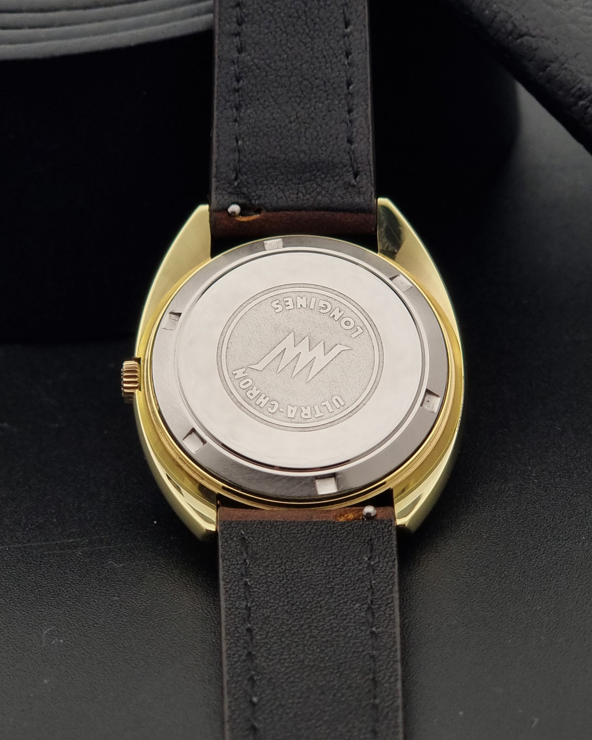 Close-up of the back of a A vintage Longines Ultra-Chron automatic watch with a gold-tone case, silver-tone screw-top back cover engraved with the Longines logo, and a dark brown leather strap on a dark background.
