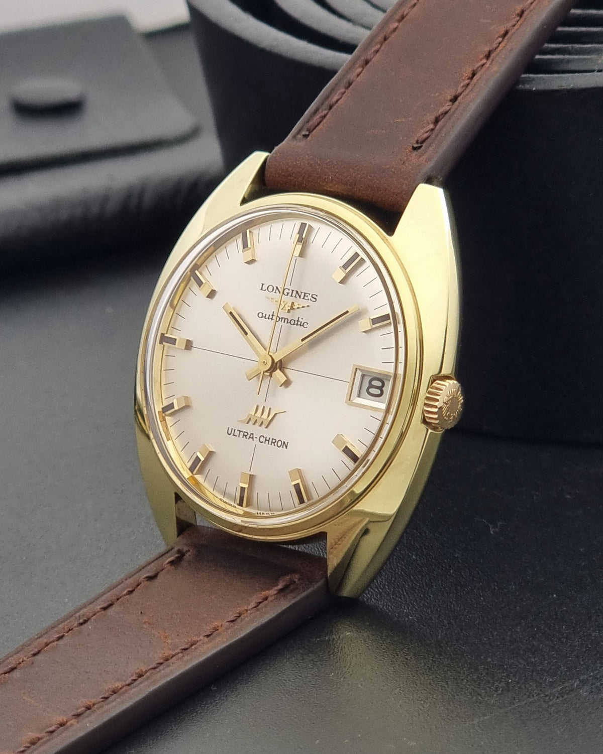 A vintage Longines automatic wristwatch with a gold-tone case, cream-colored dial, and a brown leather strap, displayed on a dark textured surface.