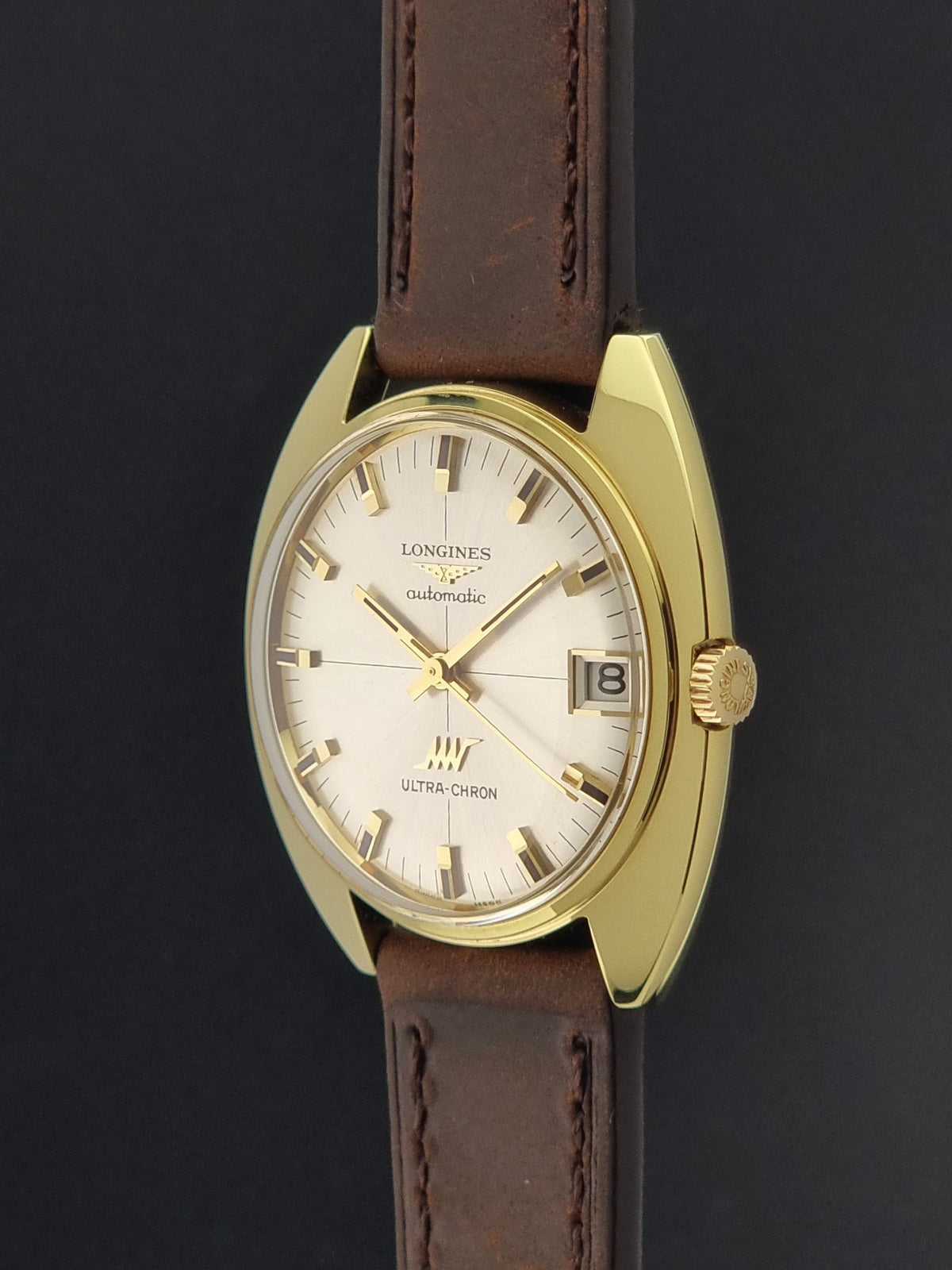 A vintage Longines Ultra-Chron watch with a gold-tone case, cream-colored dial, and a brown leather strap, shown against a dark background.