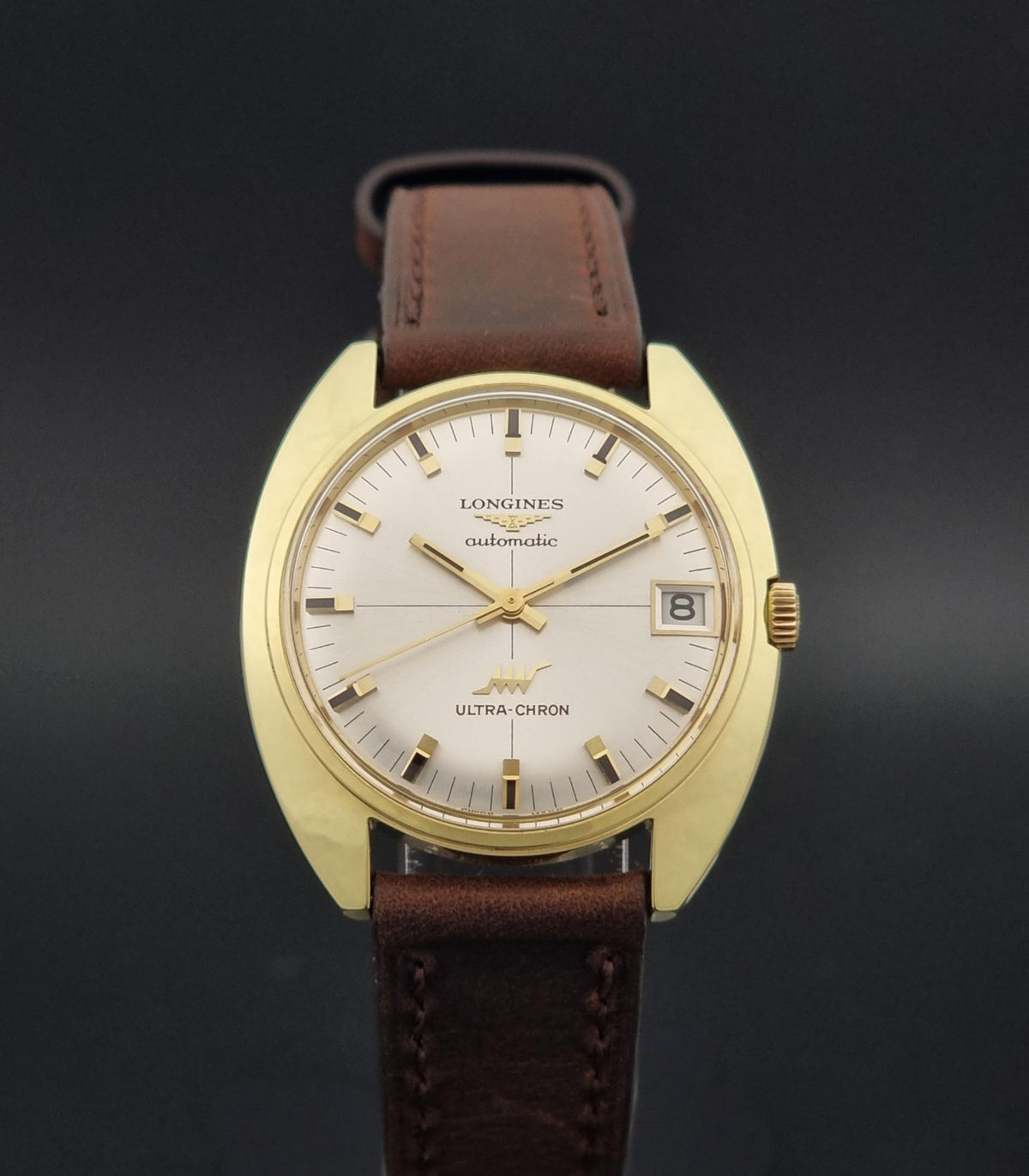 A vintage Longines Ultra-Chron watch with a gold-tone case, white dial, and brown leather strap, shown against a dark gray background.