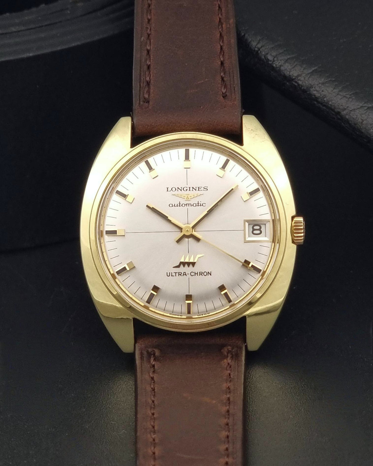 A vintage Longines Ultra-Chron automatic watch with a gold-tone case, white dial, and brown leather strap, displayed on a dark textured surface.