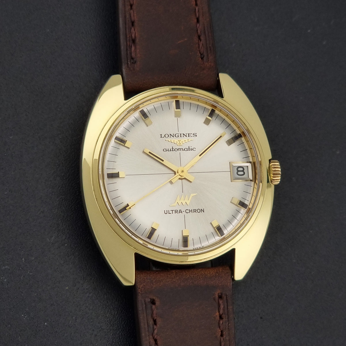 A vintage Longines Ultra-Chron automatic watch with a gold-tone case, white dial, and brown leather strap, shown against a dark textured background.