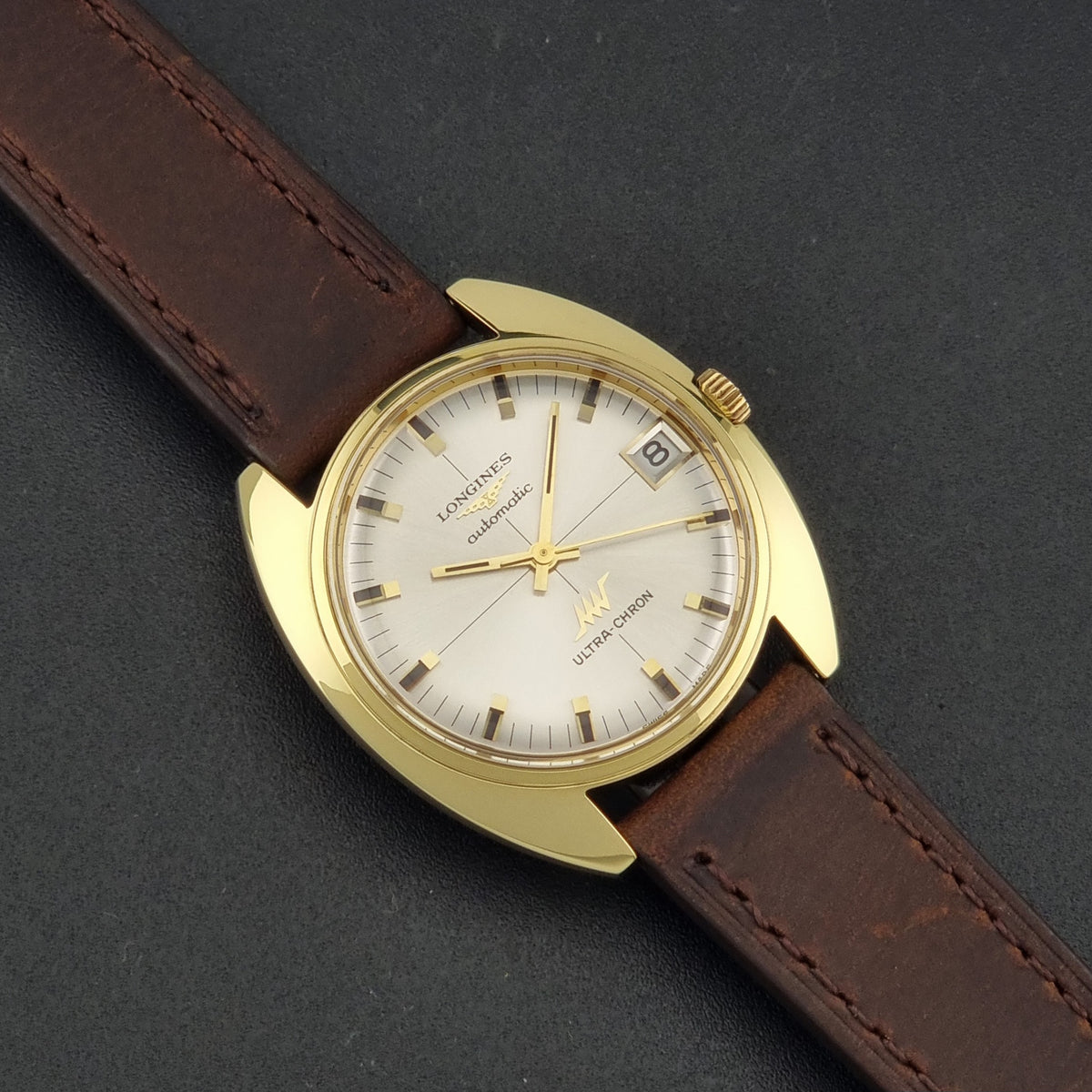 A vintage Longines watch with a gold-tone case, white dial, and brown leather strap, photographed diagonally on a dark textured background.