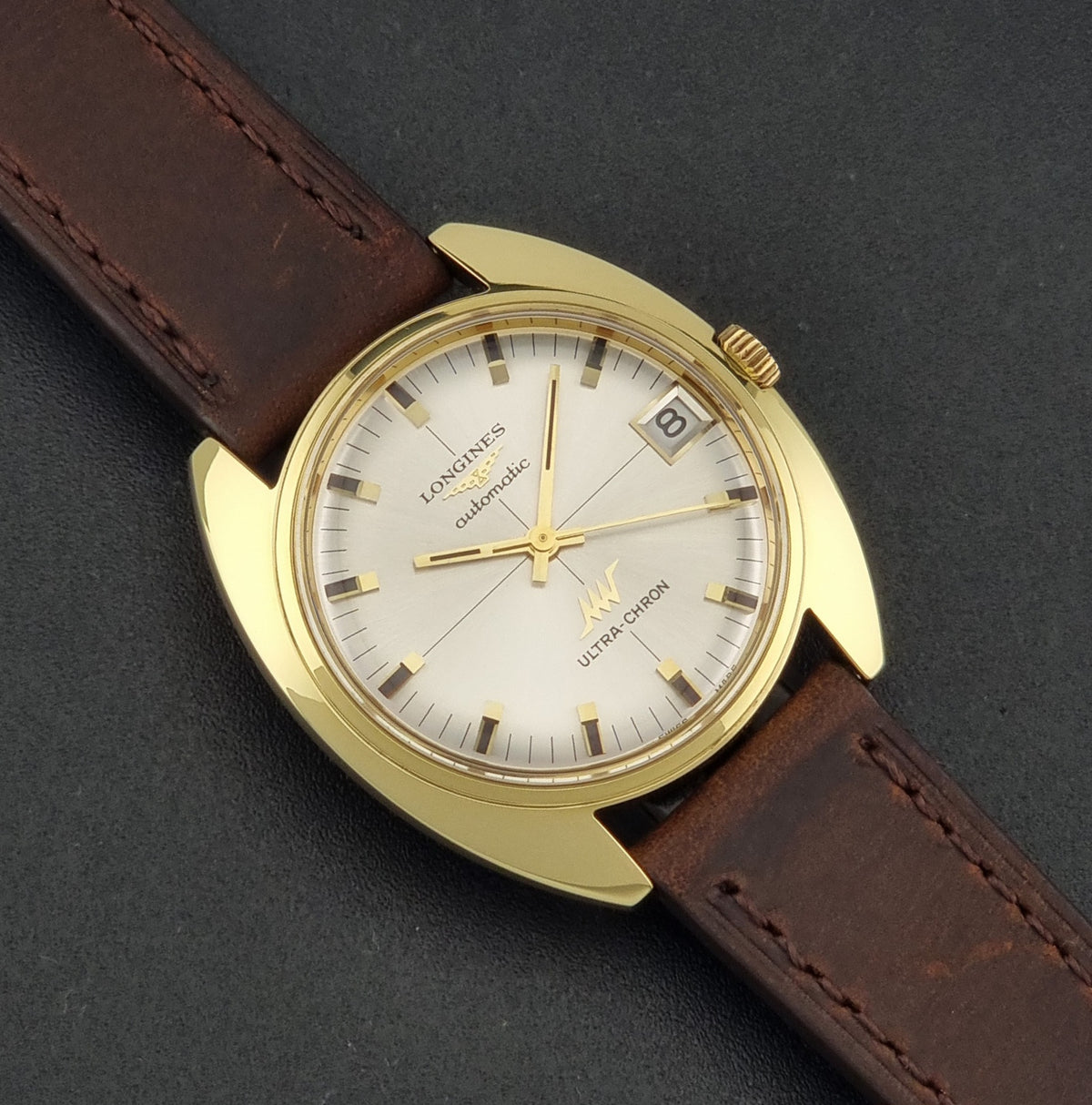 Vintage Longines ULTRA-CHRON 8073-2 watch with a gold-tone case, white dial, and brown leather strap, photographed diagonally on a dark textured background.
