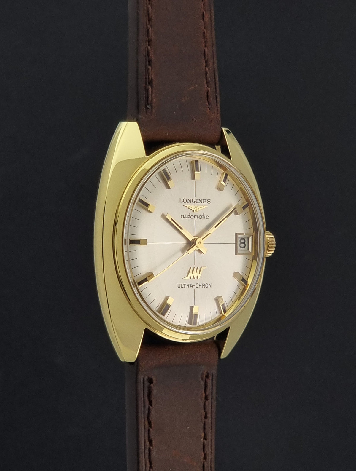 A vintage Longines watch with a gold-tone case, cream-colored dial, and a brown leather strap, shown against a dark background.