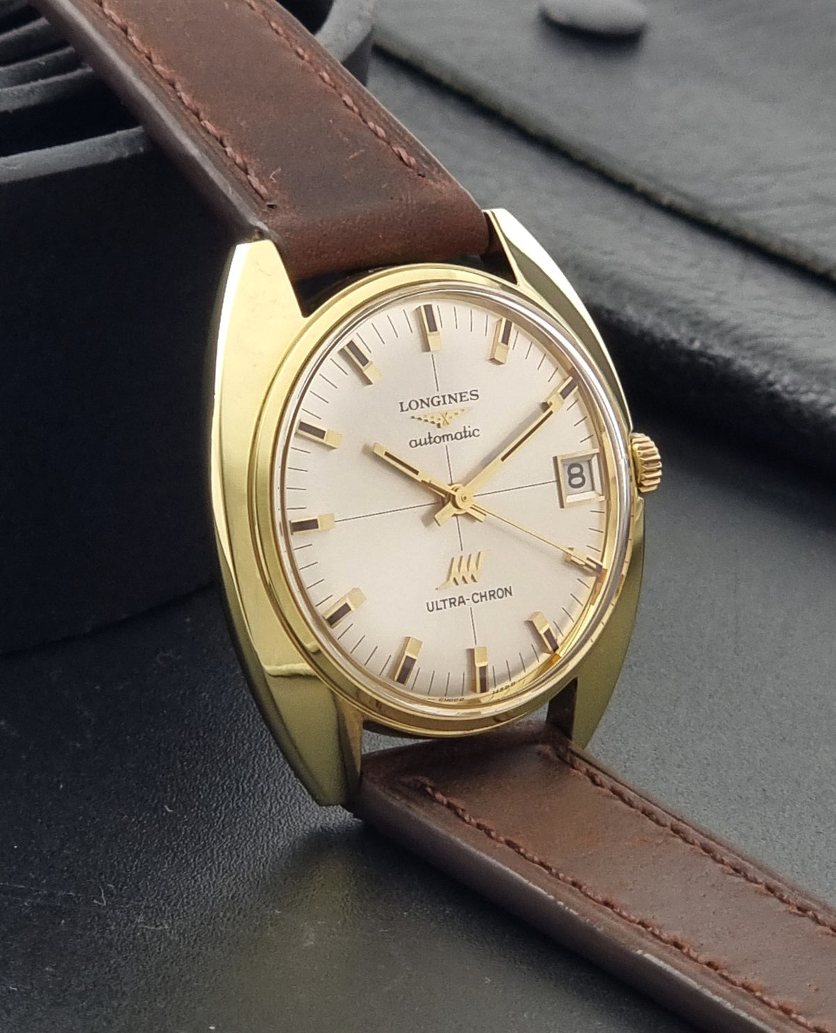 A vintage Longines Ultra Chron automatic watch with a gold-tone case, cream dial, and brown leather strap, resting on a dark textured surface.