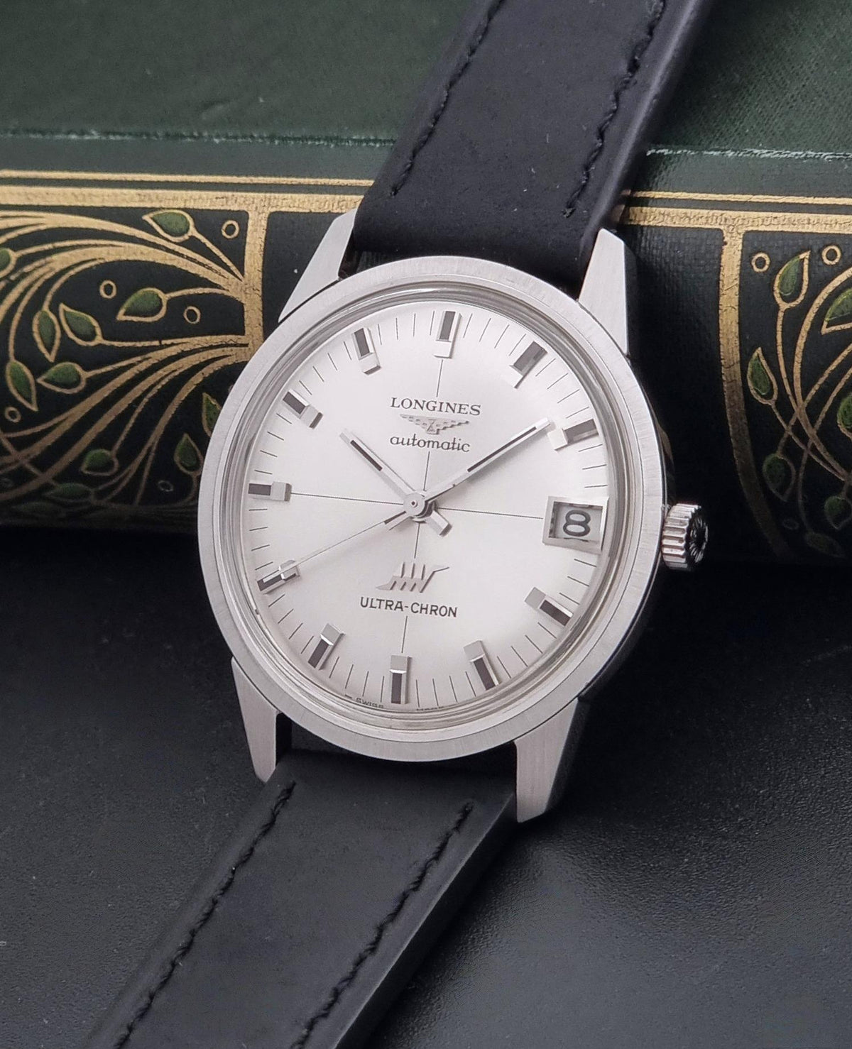 A vintage Longines Ultra-Chron watch with a silver-toned case, white dial, and black leather strap, displayed on a dark textured surface with an ornate green book in the background.