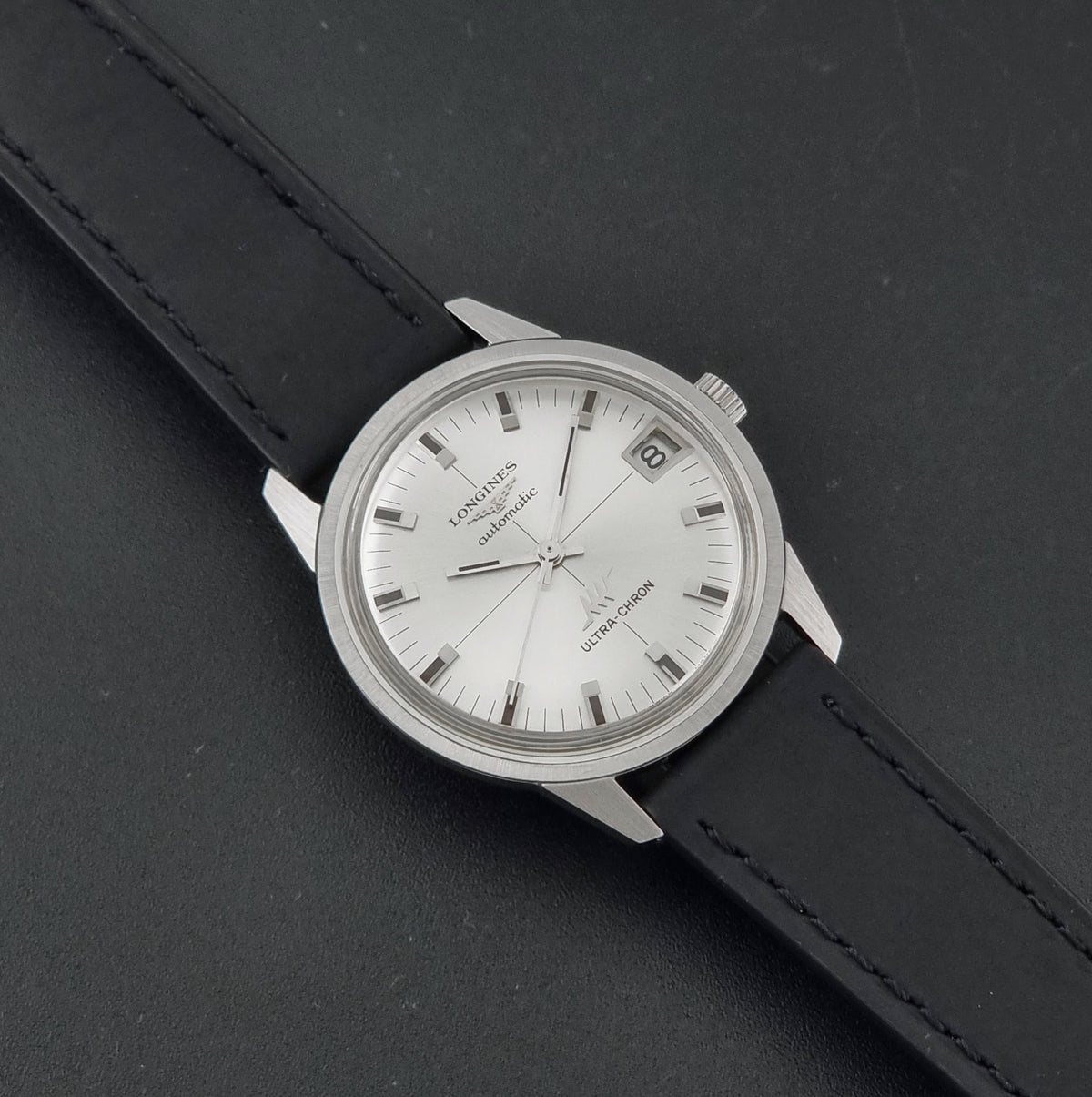 Vintage Longines ULTRA-CHRON 8309 watch with a silver-toned metal case, white dial, and black leather strap, photographed diagonally on a dark textured background.