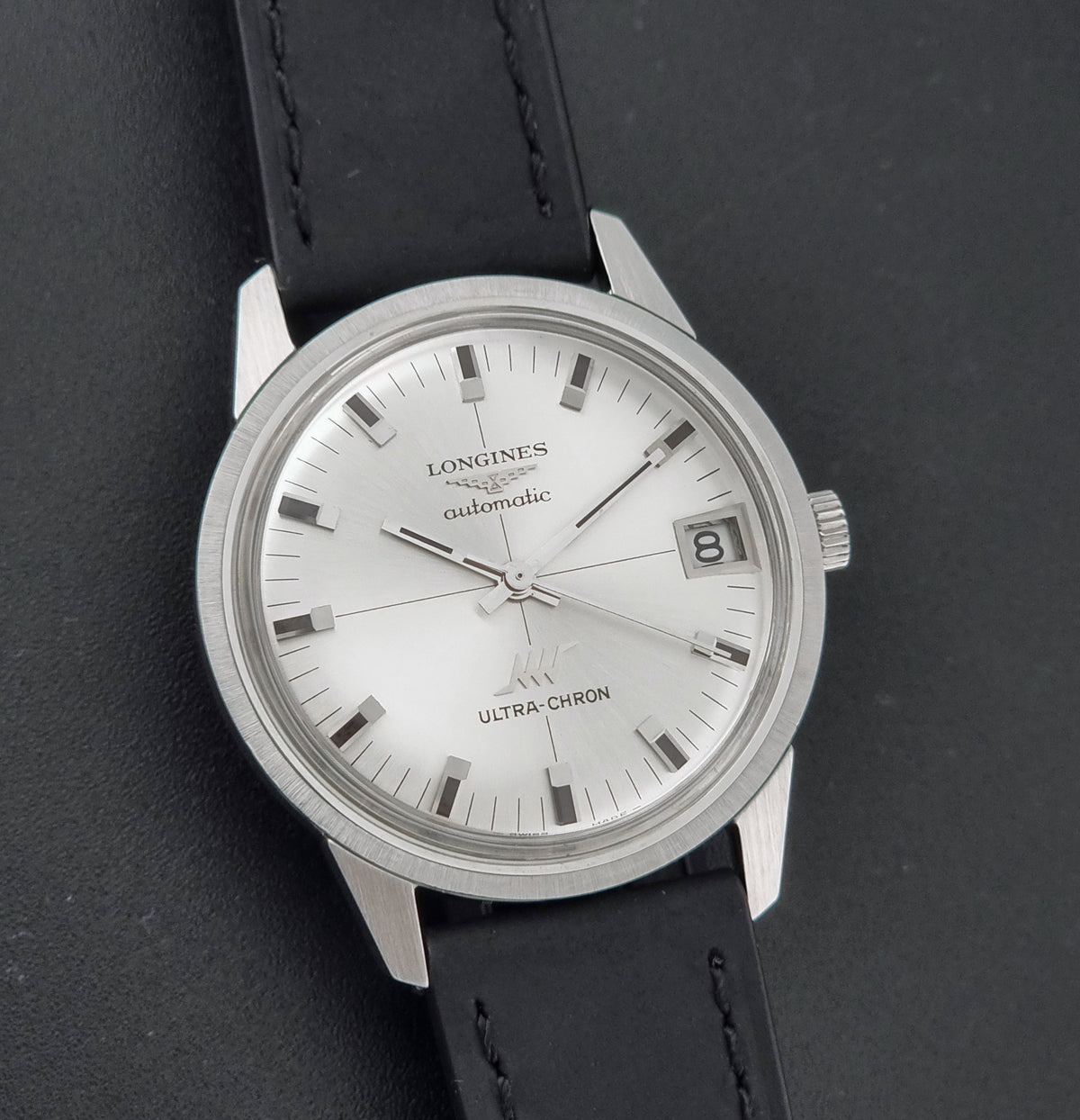 A vintage Longines automatic wristwatch with a silver-toned round case, white dial, date window, and black leather strap, photographed on a dark textured background.