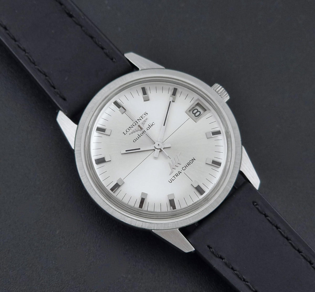 A close-up of a Vintage Longines ULTRA-CHRON 8309 watch with a silver-toned stainless steel case, silver dial, and black leather strap on a dark textured background.
