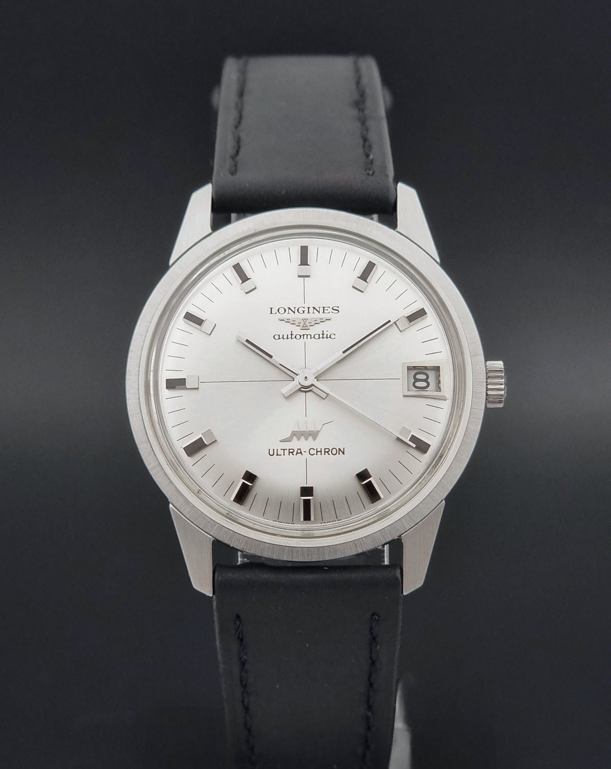 A Longines Ultra-Chron automatic watch with a silver-toned stainless steel case, white dial, and black leather strap, shown against a dark gray background.
