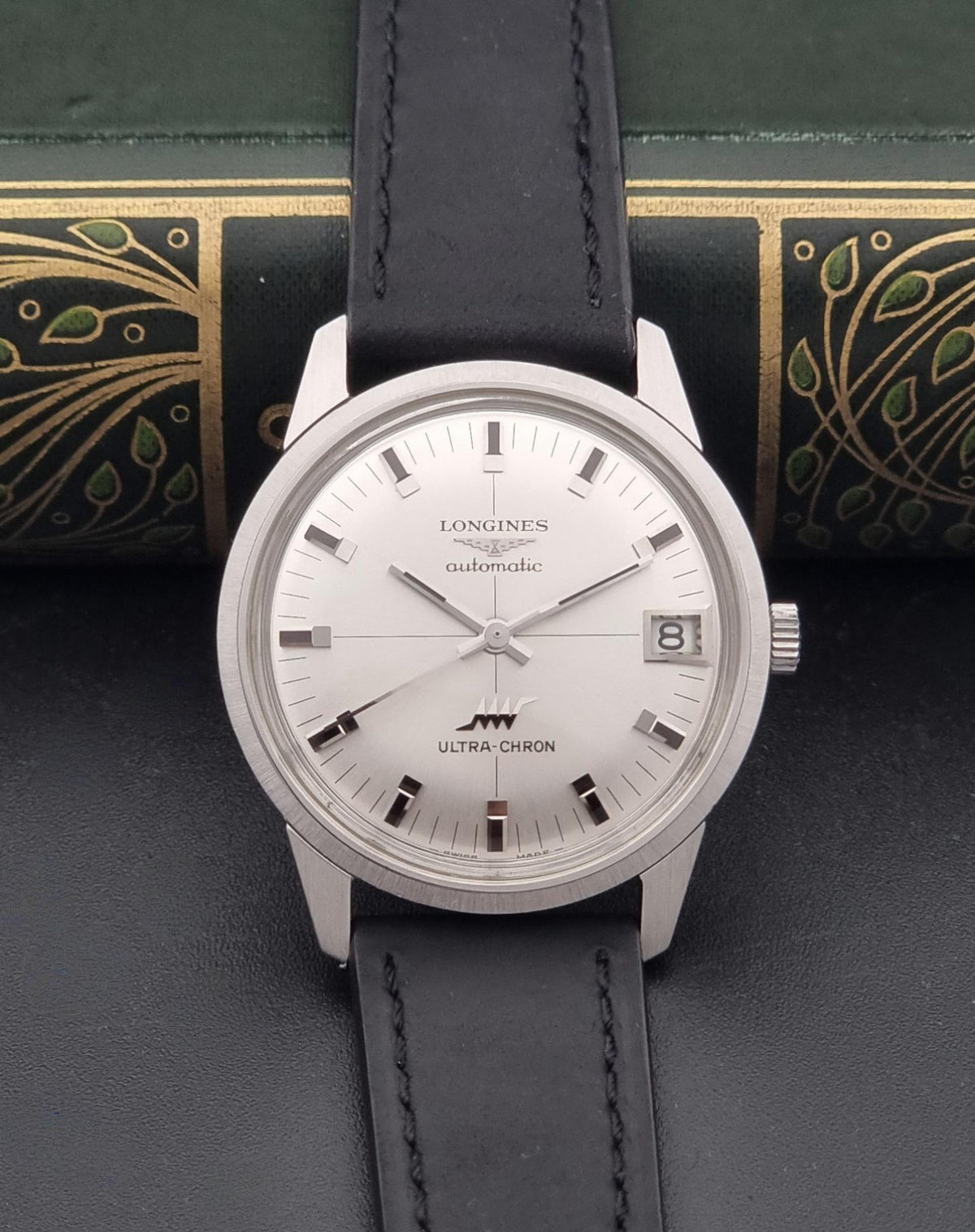 A vintage Longines watch with a silver-toned stainless steel case, white dial, and black leather strap, displayed against a dark textured background with an ornate green book spine behind it.