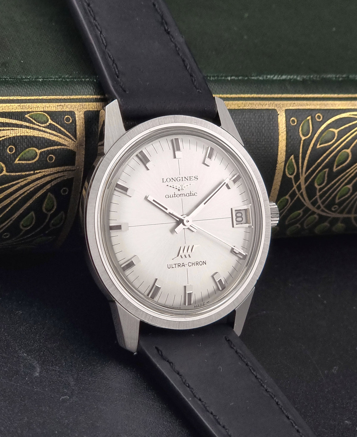 A Longines UltraChron automatic watch with a silver-toned stainless steel case, white dial, and black leather strap, displayed diagonally on a dark textured surface with an antique-style green and gold book in the background.