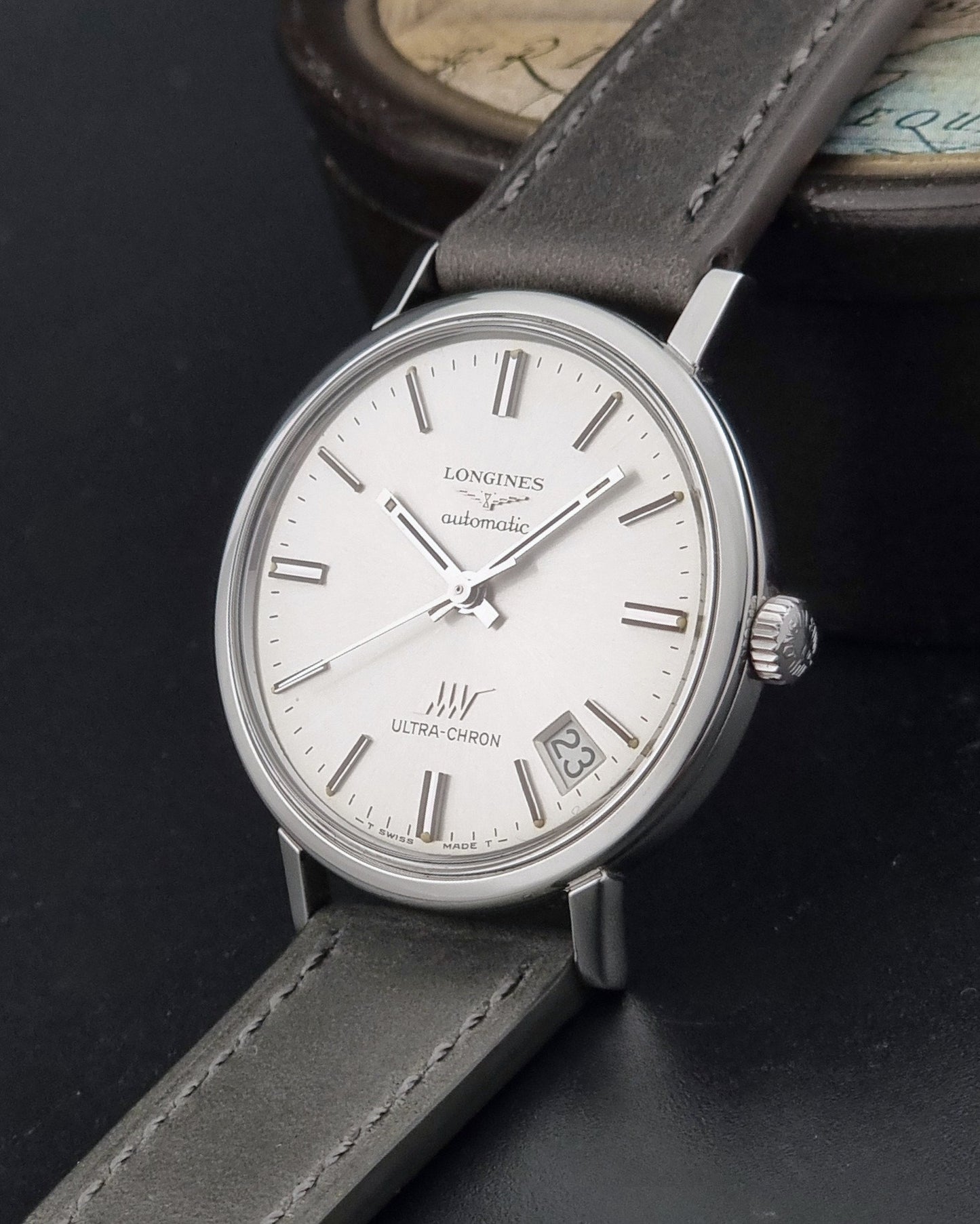 Vintage Longines ULTRA-CHRON 8383-1 watch with a silver-toned round case and gray leather strap on a dark gray background crown view