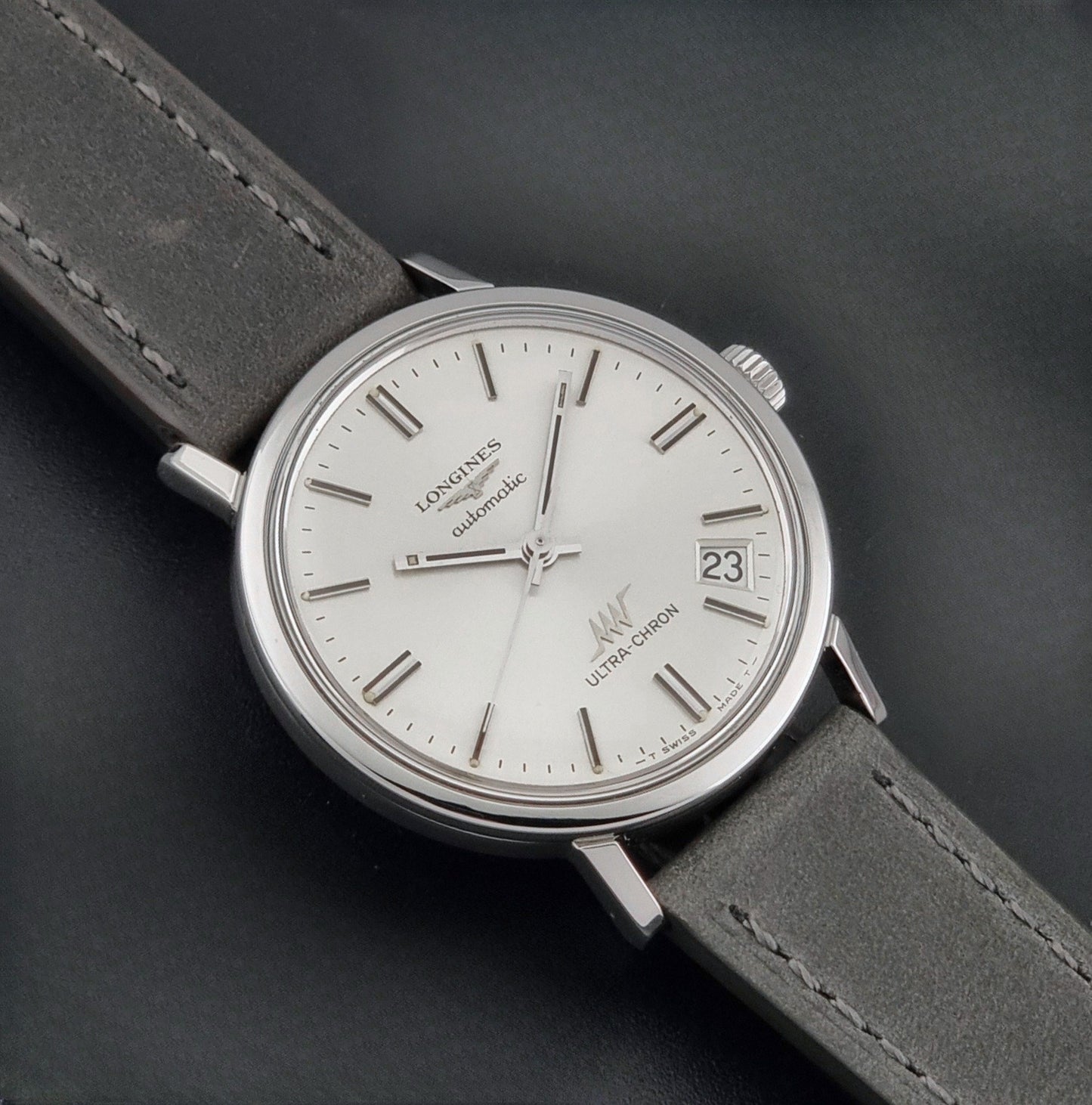 Vintage Longines ULTRA-CHRON 8383-1 watch with a silver-toned round case and gray leather strap on a dark gray background close up dial view