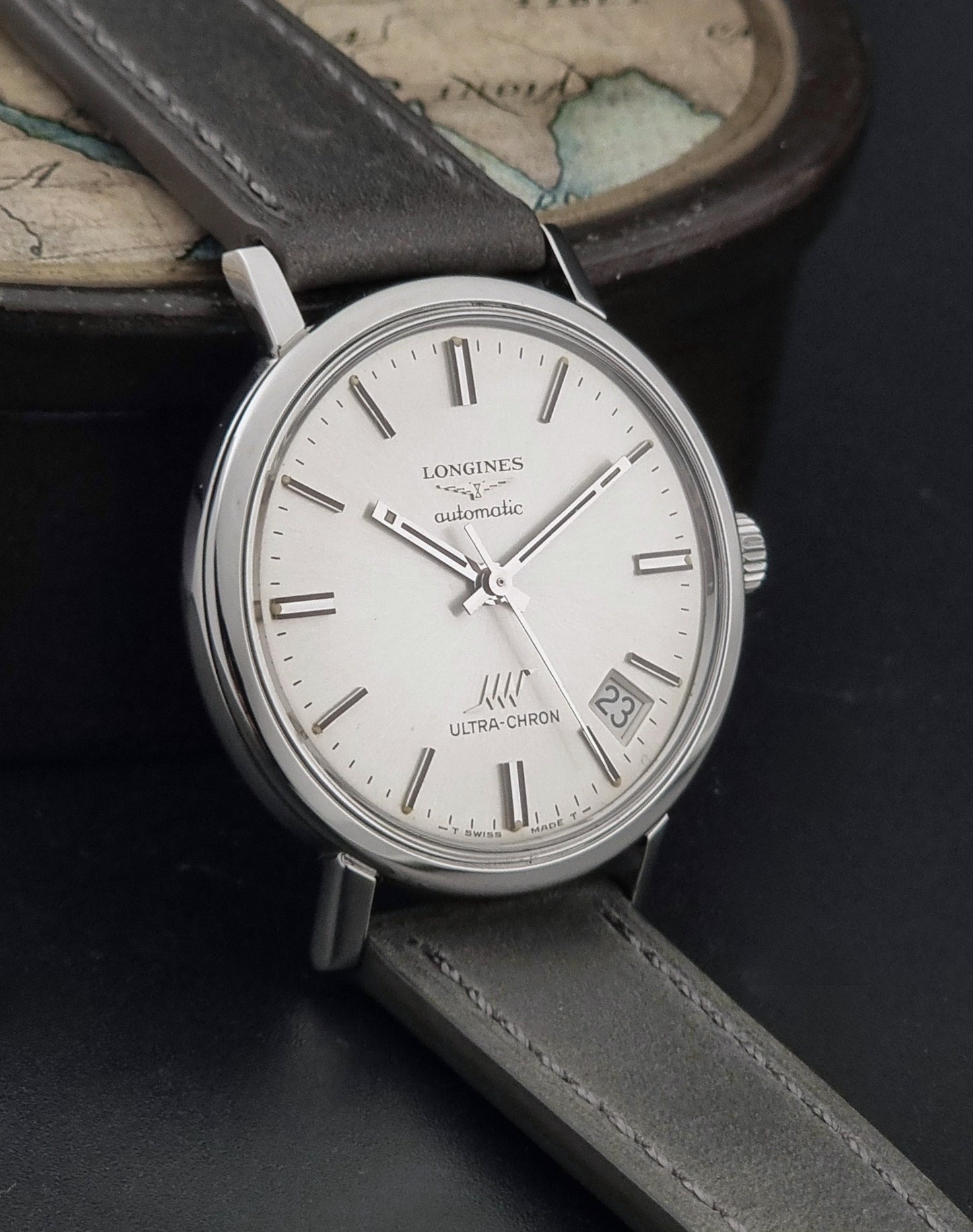 Vintage Longines ULTRA-CHRON 8383-1 watch with a silver-toned round case and gray leather strap on a dark gray background side view