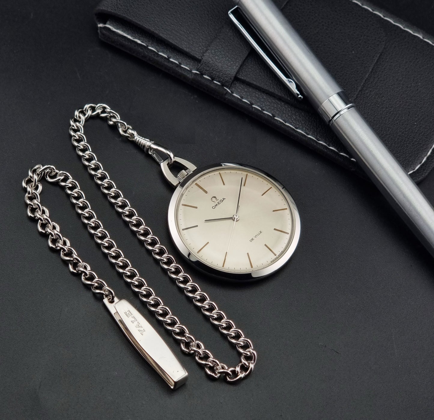 A silver-tone Omega 131.1714 pocket watch with a matching chain and rectangular tag, displayed on a dark textured surface next to a silver pen and a black leather case.
